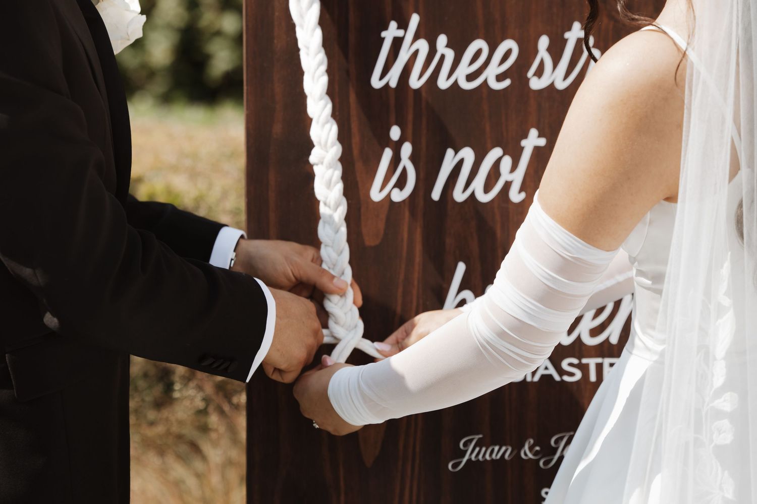 Close-up of bride and groom hands braiding white unity cord at outdoor wedding ceremony.