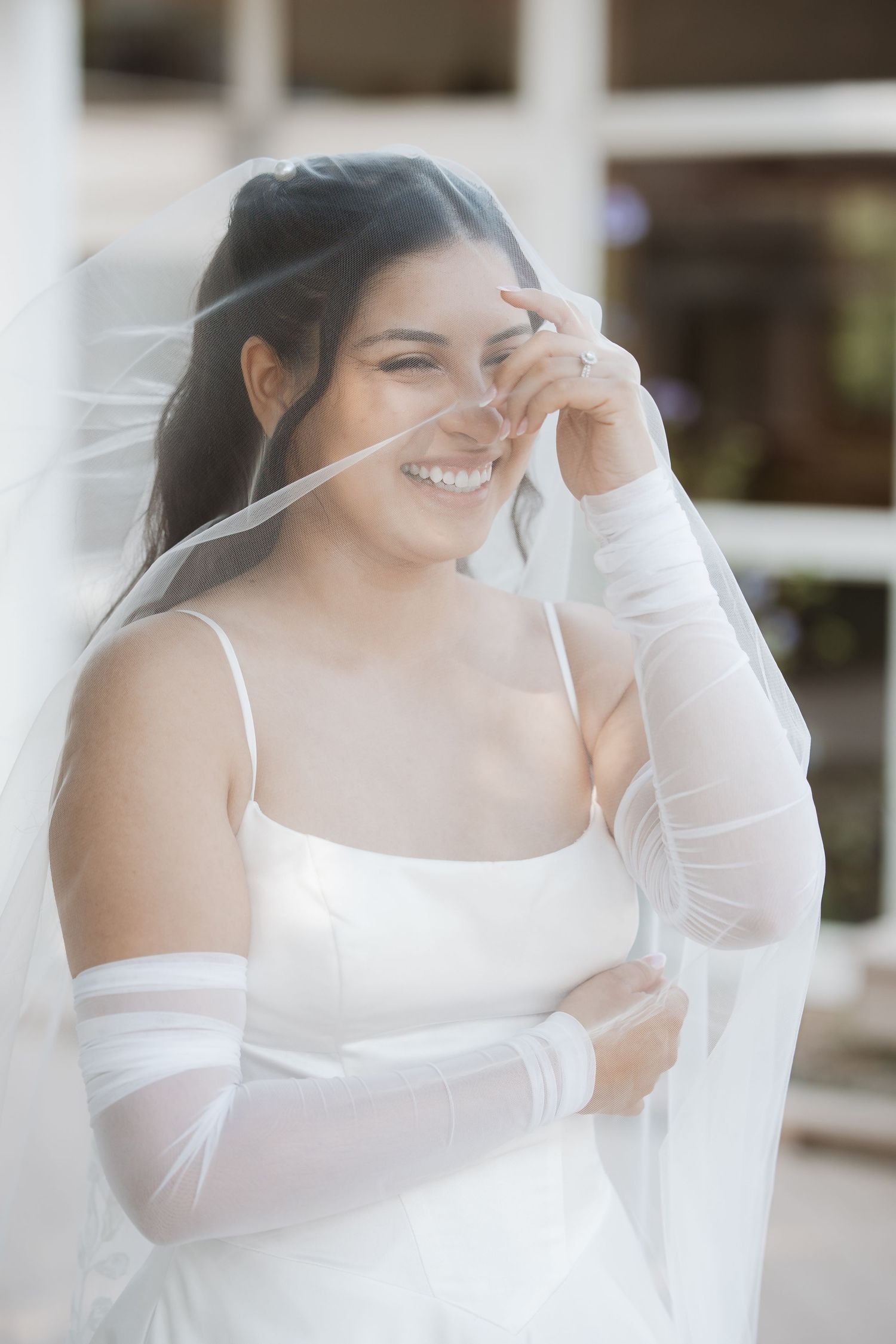 Laughing bride in white gown adjusts her flowing veil over her face.