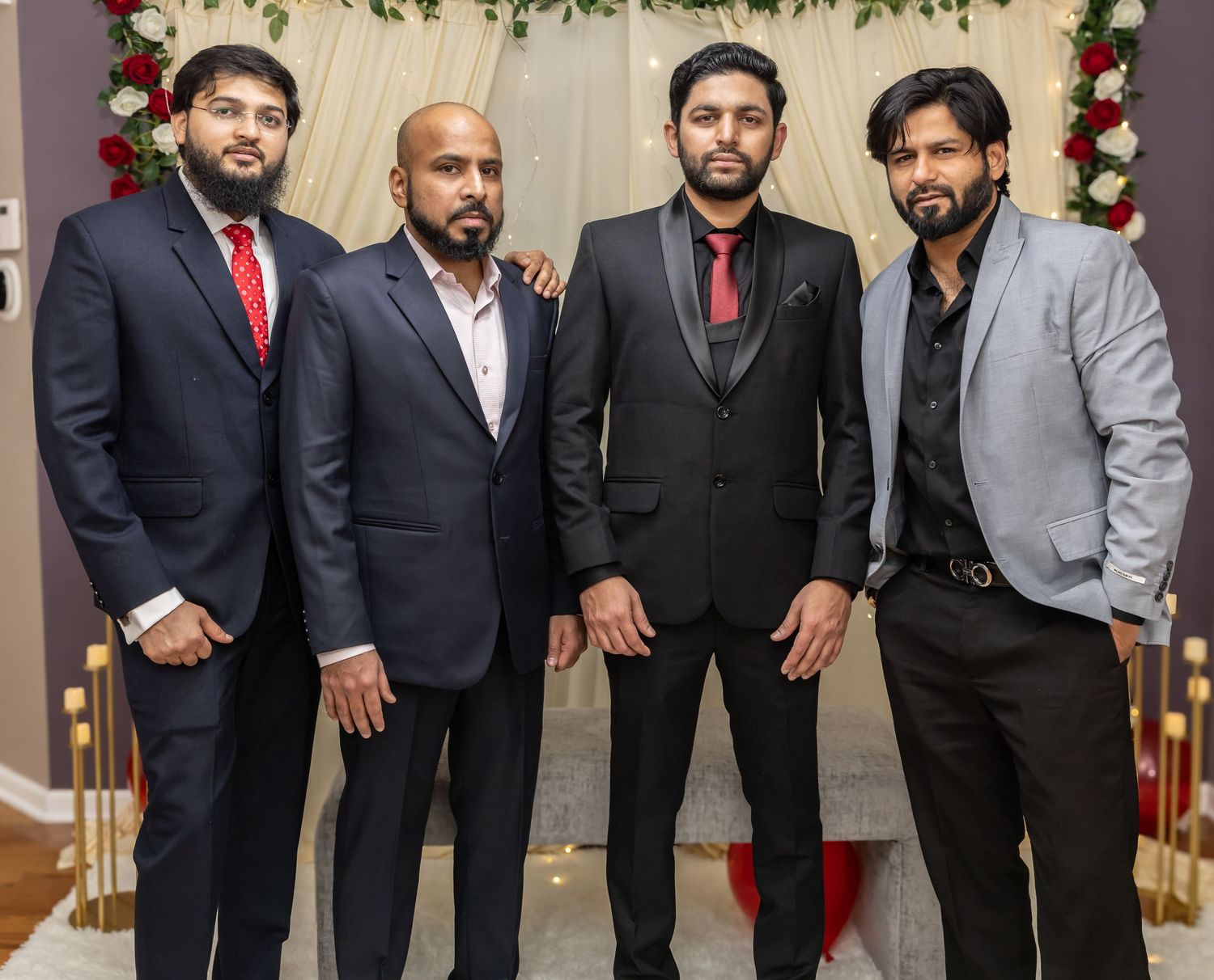 Four well-dressed men in formal suits smile together at a wedding celebration with floral decorations.