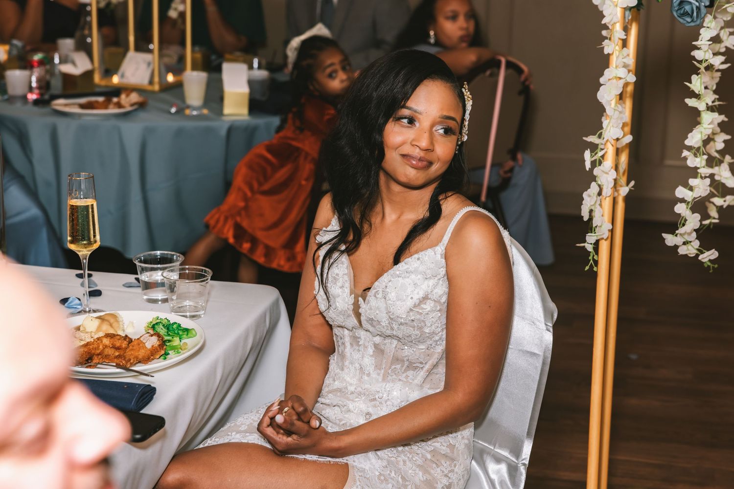Beautiful bride in lace wedding gown sitting at reception table, smiling gracefully at wedding celebration.