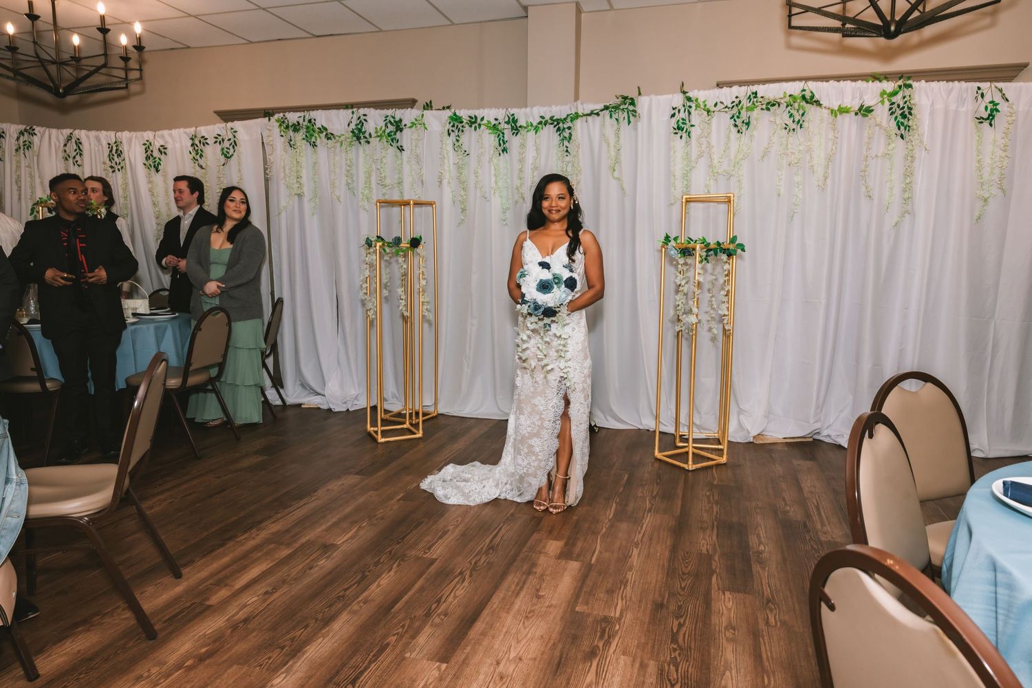 Bride in lace wedding gown holds bouquet while standing in decorated wedding reception venue.