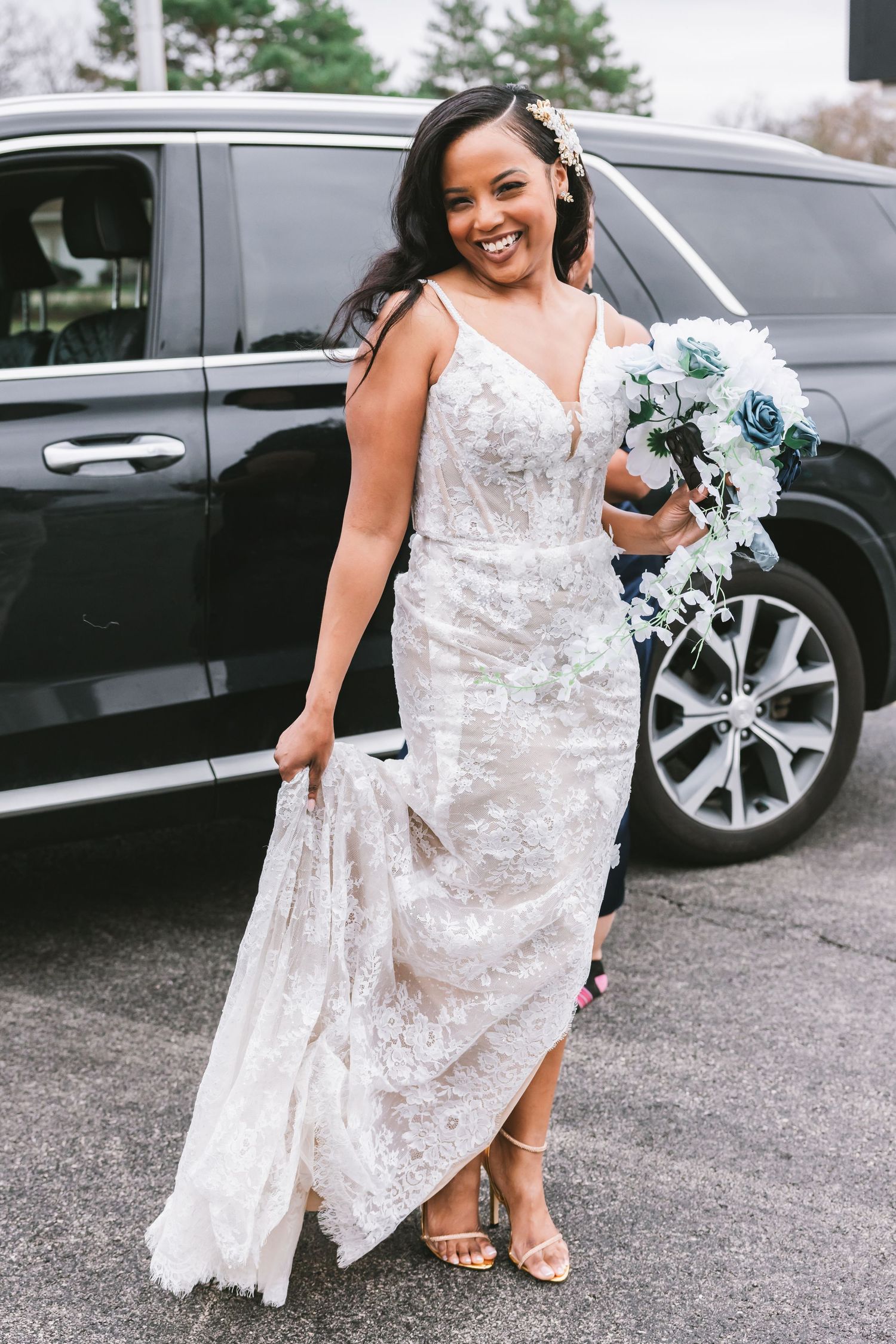 Smiling bride in lace wedding gown holds blue and white bouquet outside near a black SUV.