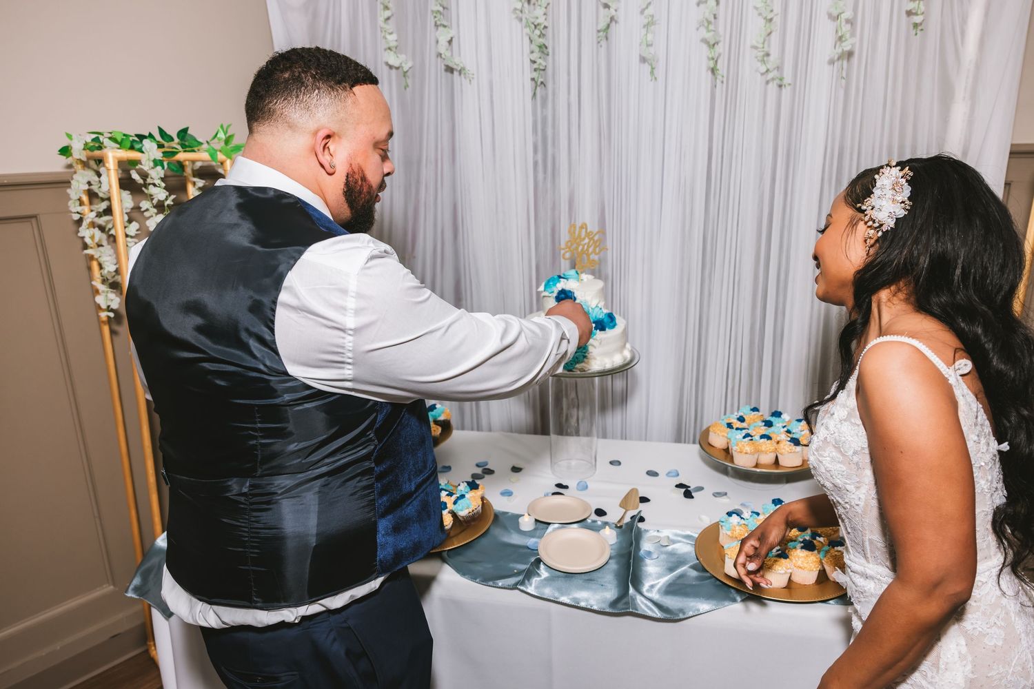 Bride and groom cutting wedding cake together at reception table with blue decorative accents.