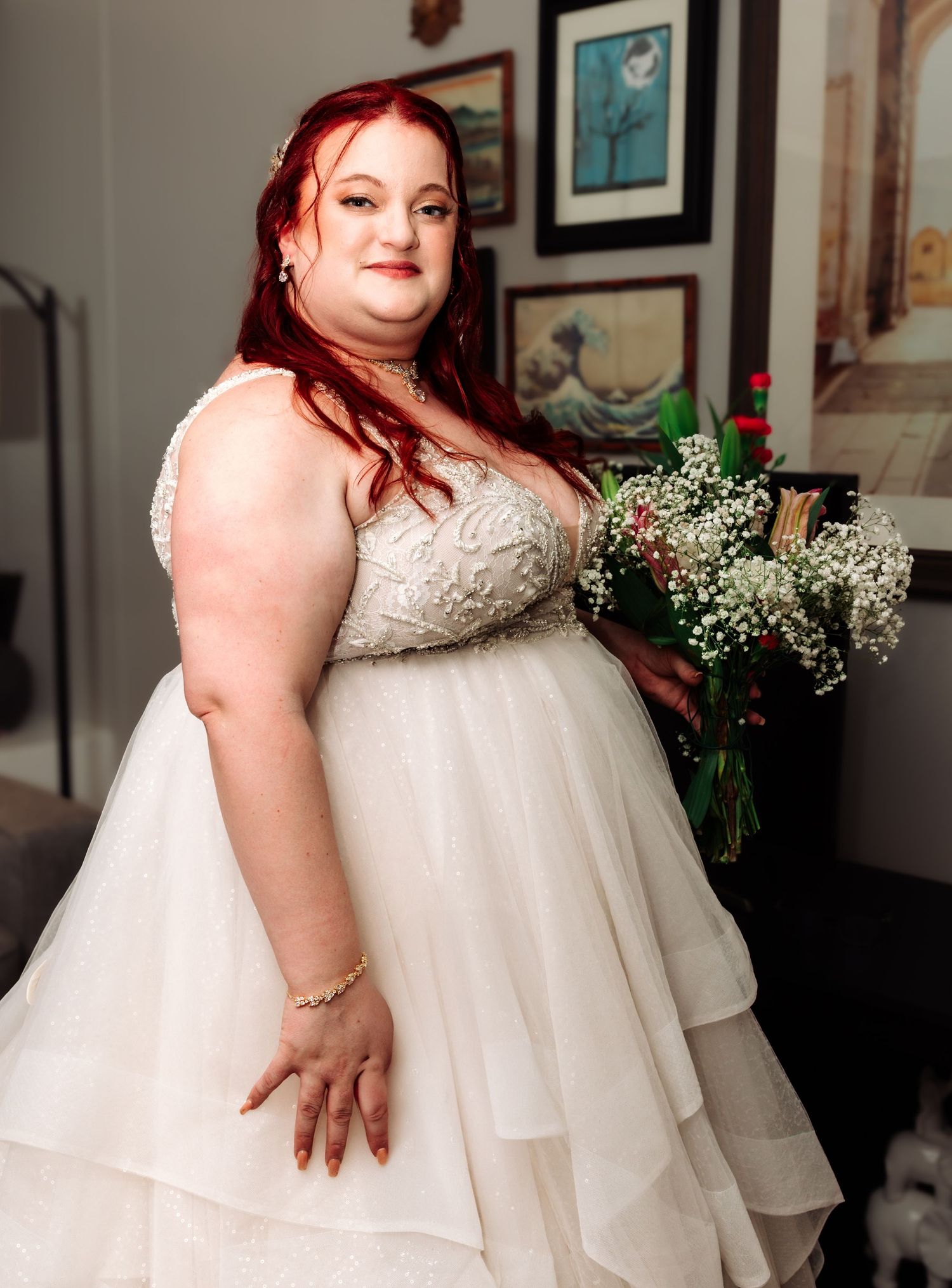 A plus-size bride with red hair in a beaded white ballgown smiles while holding a floral bouquet.