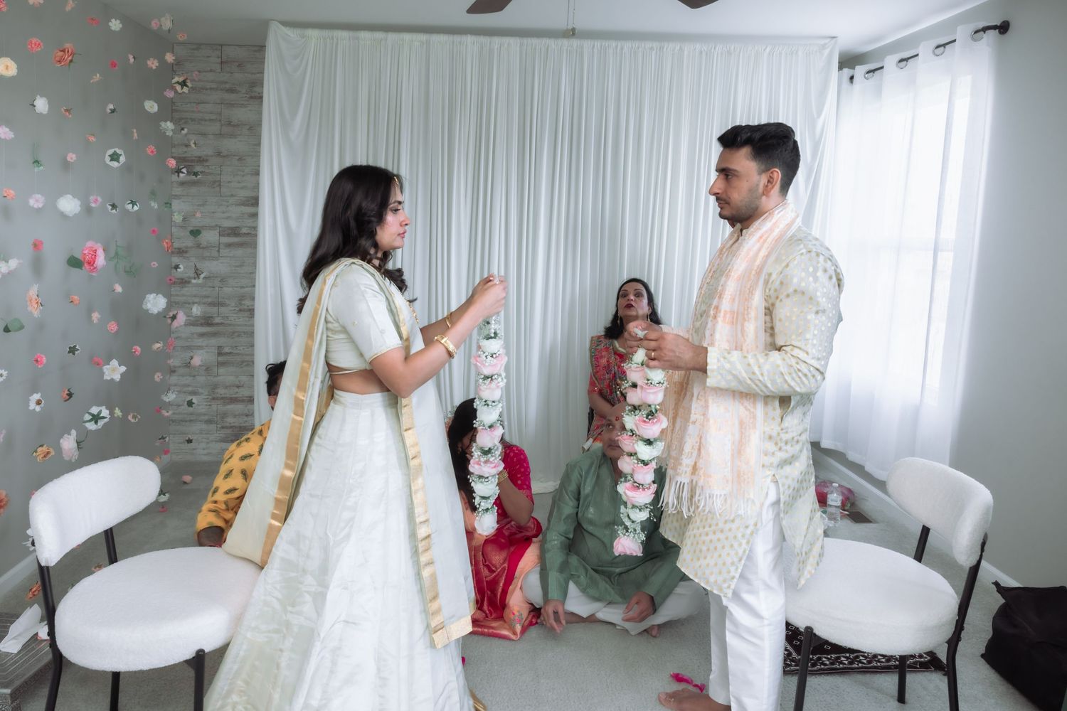 A bride in white lehenga and groom in sherwani exchange garlands during an Indian wedding ceremony.