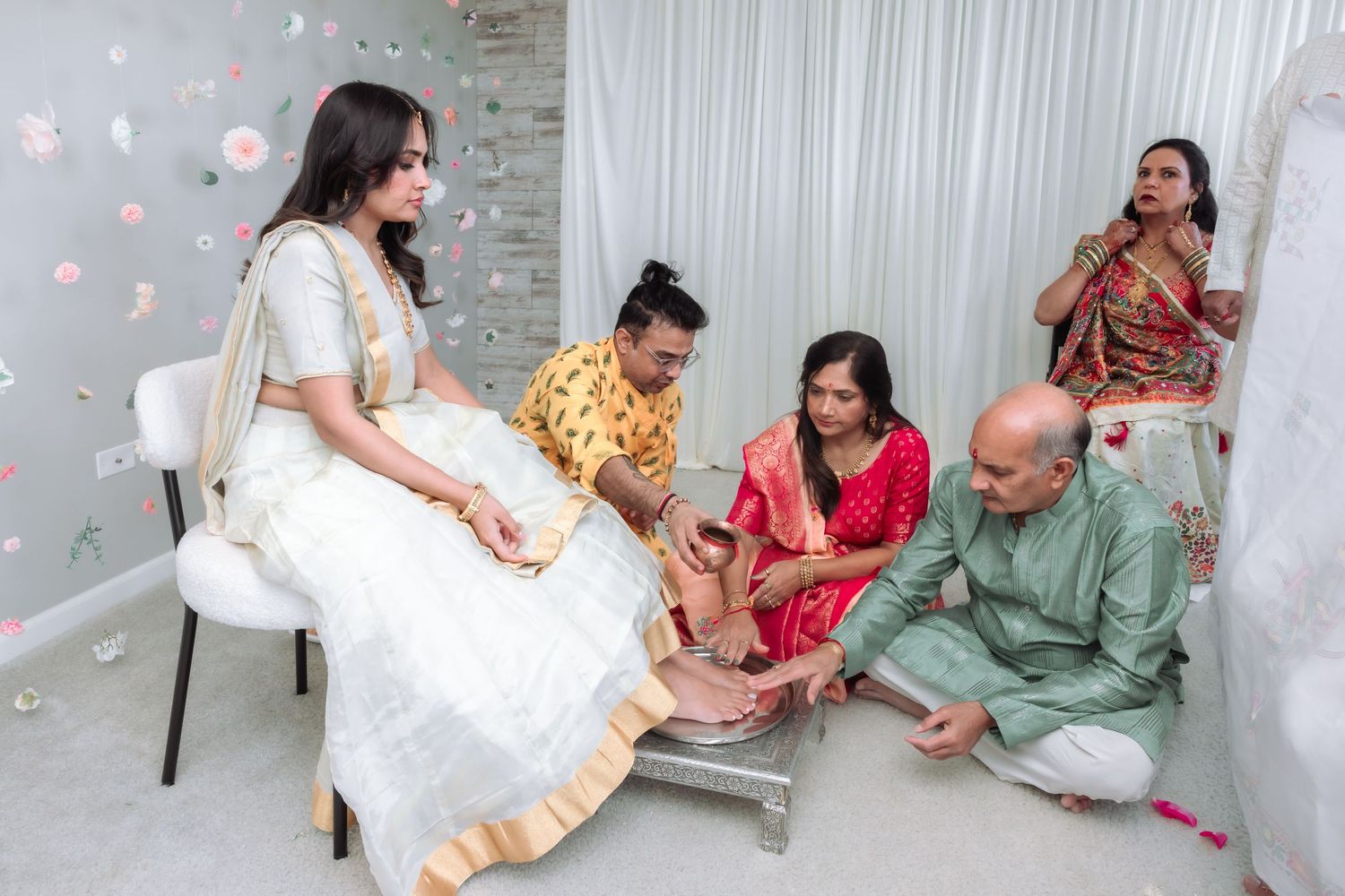 Family performs traditional Indian wedding foot ritual with bride in white lehenga surrounded by relatives.