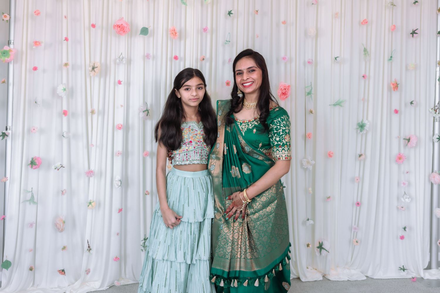 Two women in elegant green Indian attire pose together at a festive celebration with floral backdrop.
