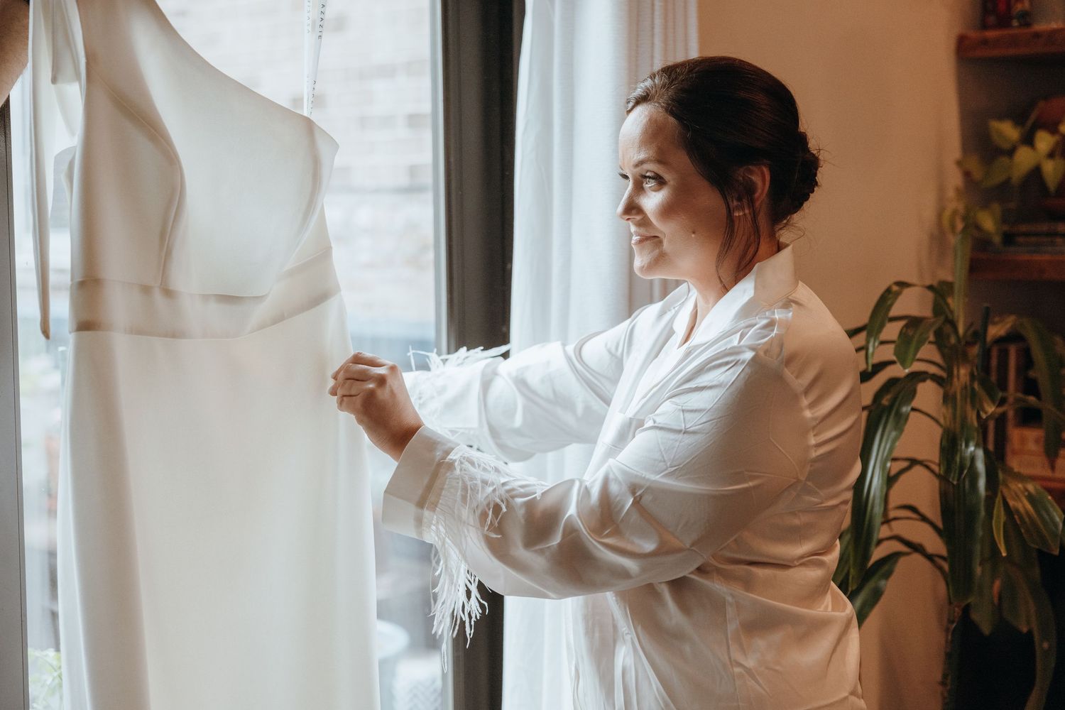 Bride in white feather-trimmed robe examining her elegant white wedding gown by a bright window.