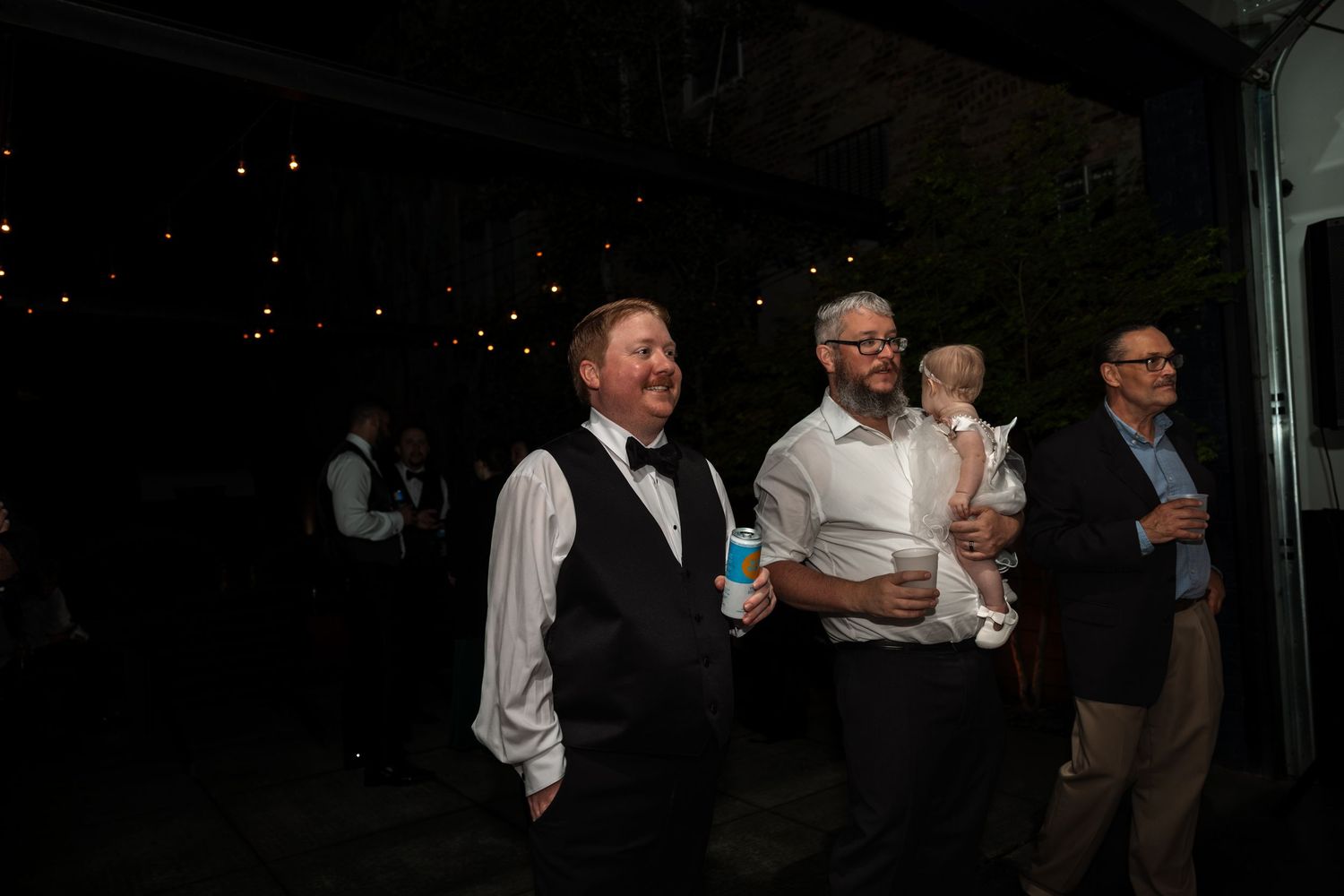 Guests in formal attire smile and socialize outdoors at evening wedding reception celebration.