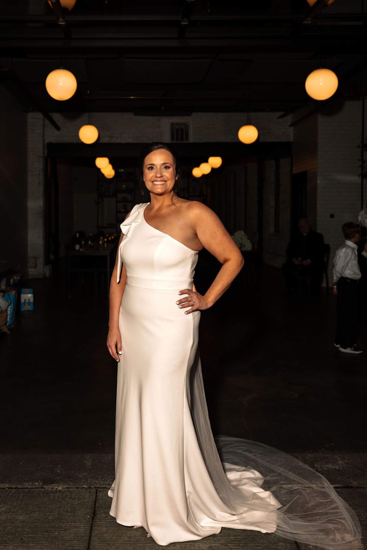 Smiling bride in elegant one-shoulder white gown with train poses confidently in dimly lit venue with warm globe lights.