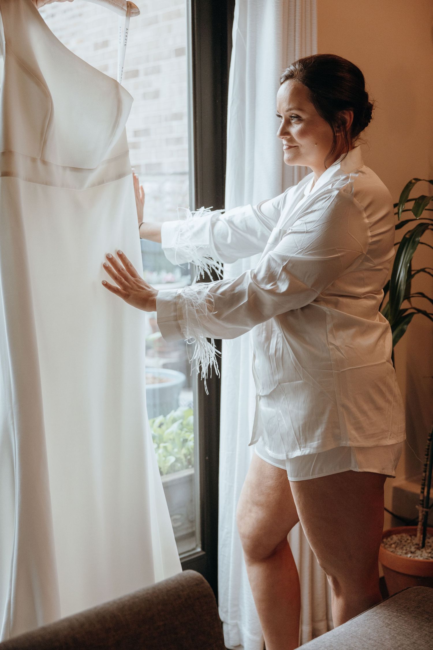 Bride in white feather-trimmed robe admiring her wedding dress hung near a sunny window.