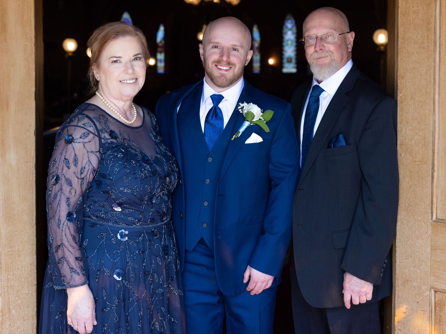 Groom in blue suit poses with parents outside church entrance before wedding ceremony.