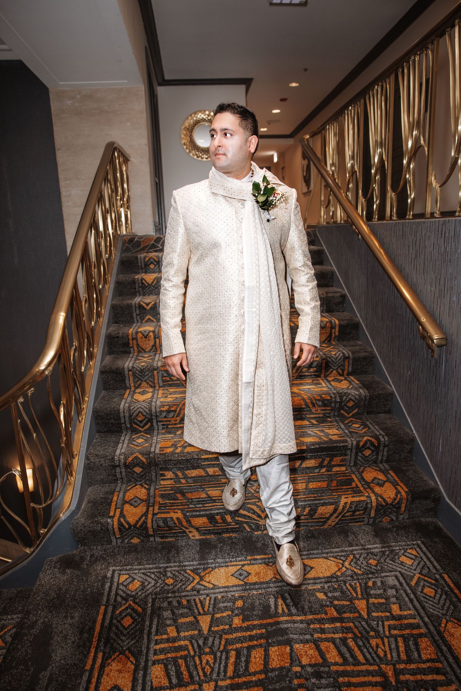 Groom in embroidered ivory sherwani with floral boutonniere walking down patterned carpeted staircase in luxurious hotel interior.