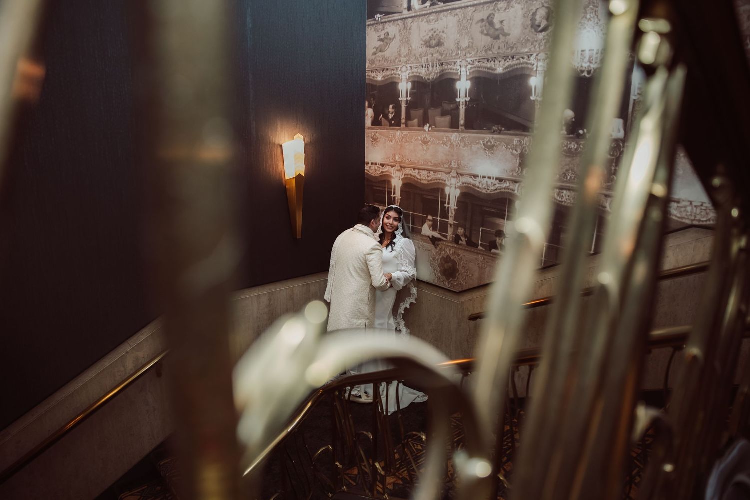 Romantic couple embracing on staircase captured through golden candelabras in warmly lit wedding venue.