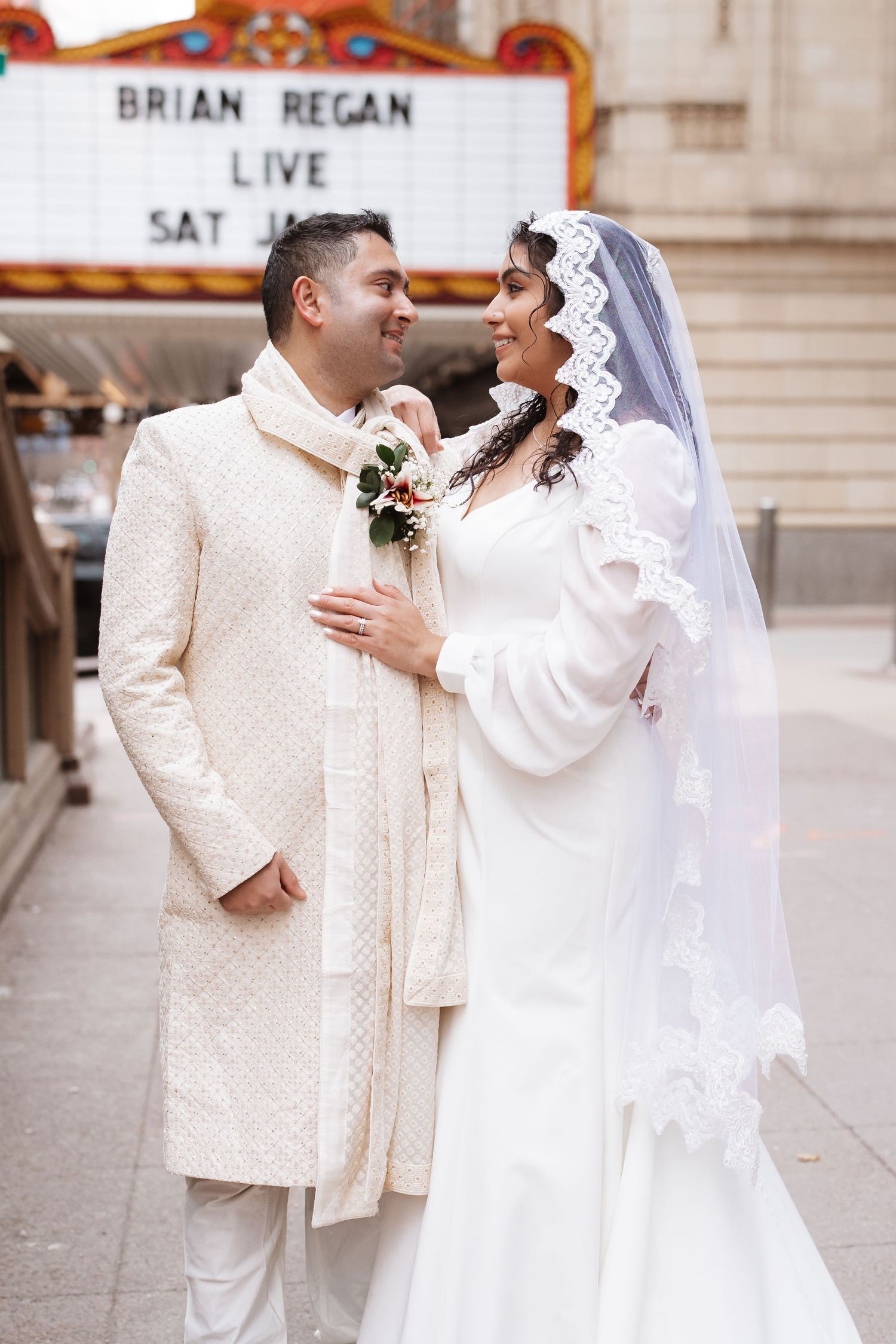South Asian couple in white wedding attire gaze at each other near Brian Regan Chicago Theatre sign.