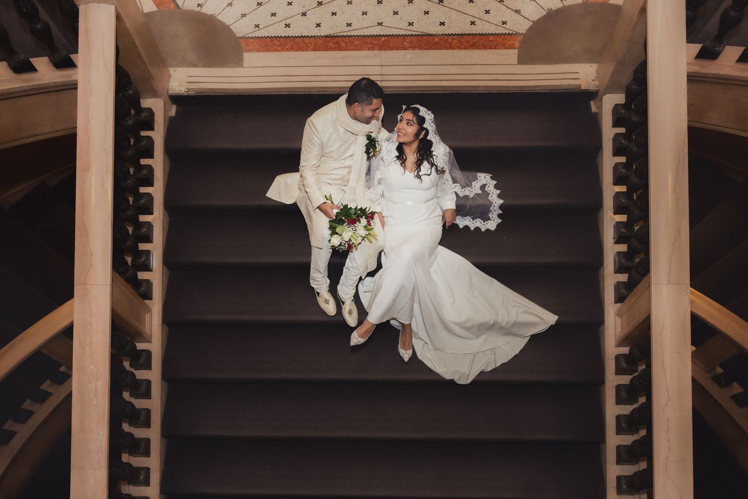 Top-down view of wedding couple seated on elegant wooden staircase, bride in flowing white dress with bouquet.