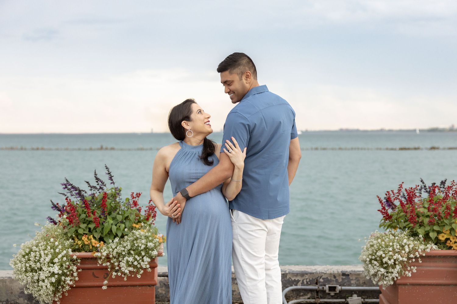 Expecting couple gazing at each other by the lake, woman in blue halter dress, man in blue shirt and white pants.