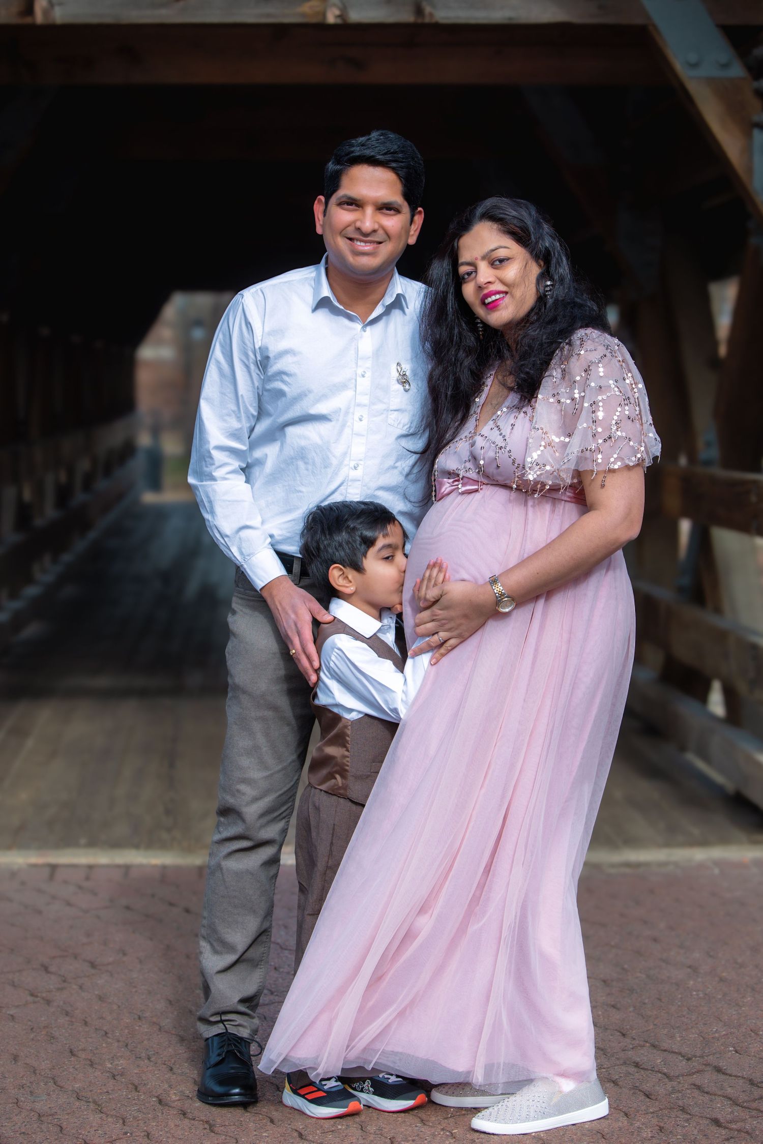 Indian family maternity portrait with father, young son, and pregnant mother in pink gown on a covered bridge.