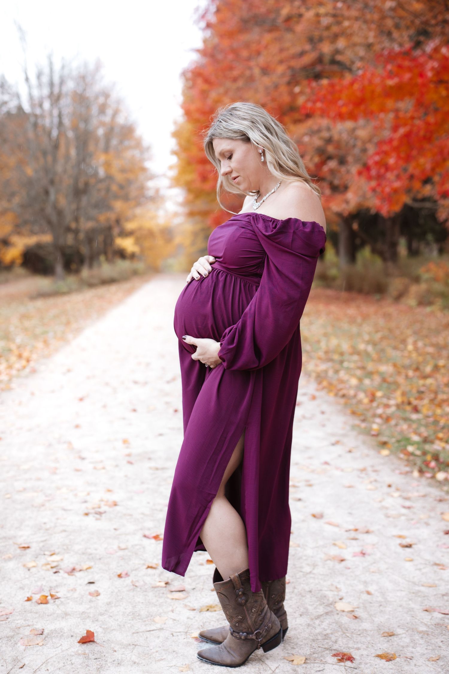 Maternity photo of pregnant woman in purple dress and boots on colorful autumn tree-lined path.