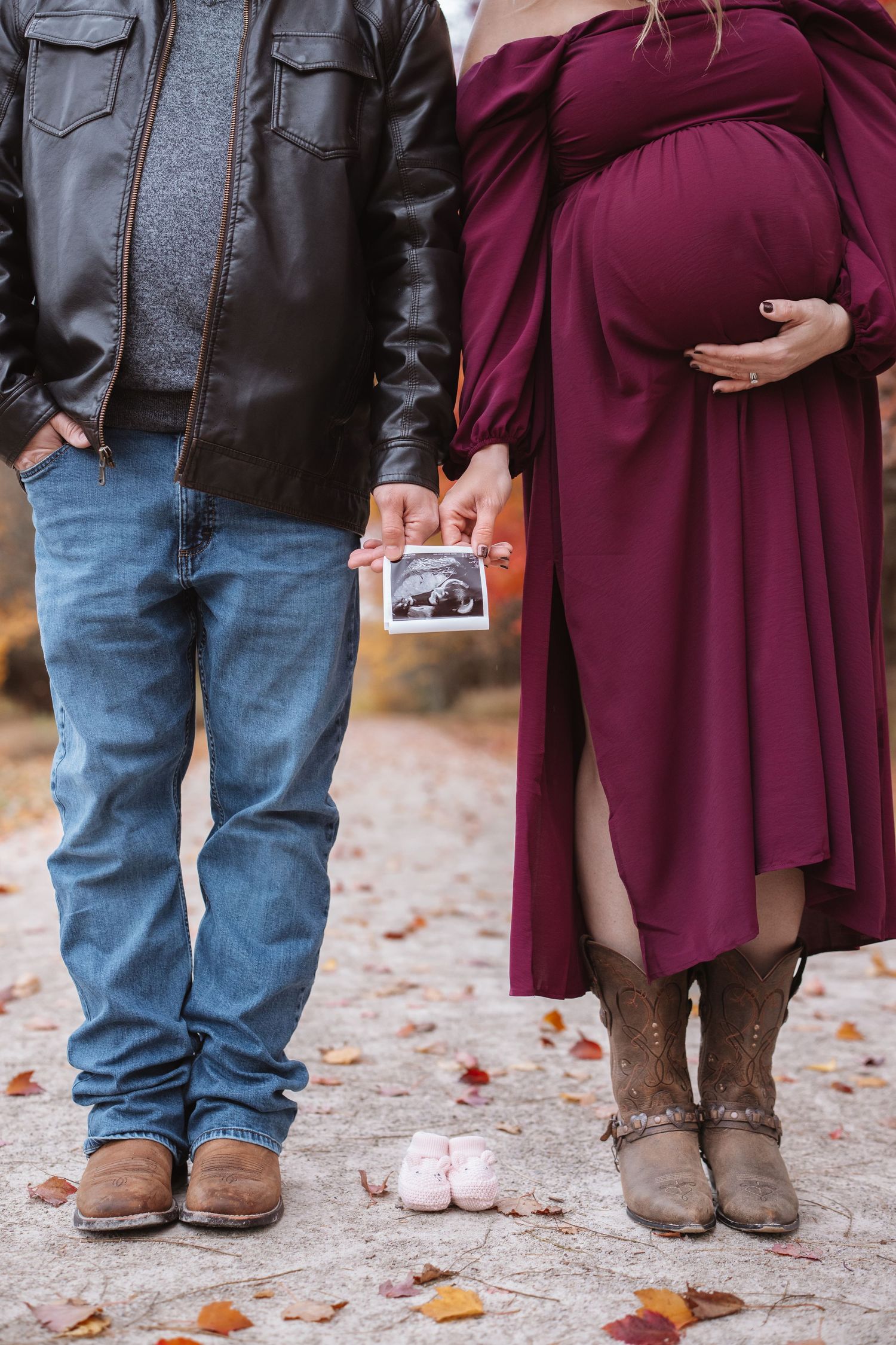 Expecting couple holding ultrasound image with pink newborn shoes placed on autumn ground.
