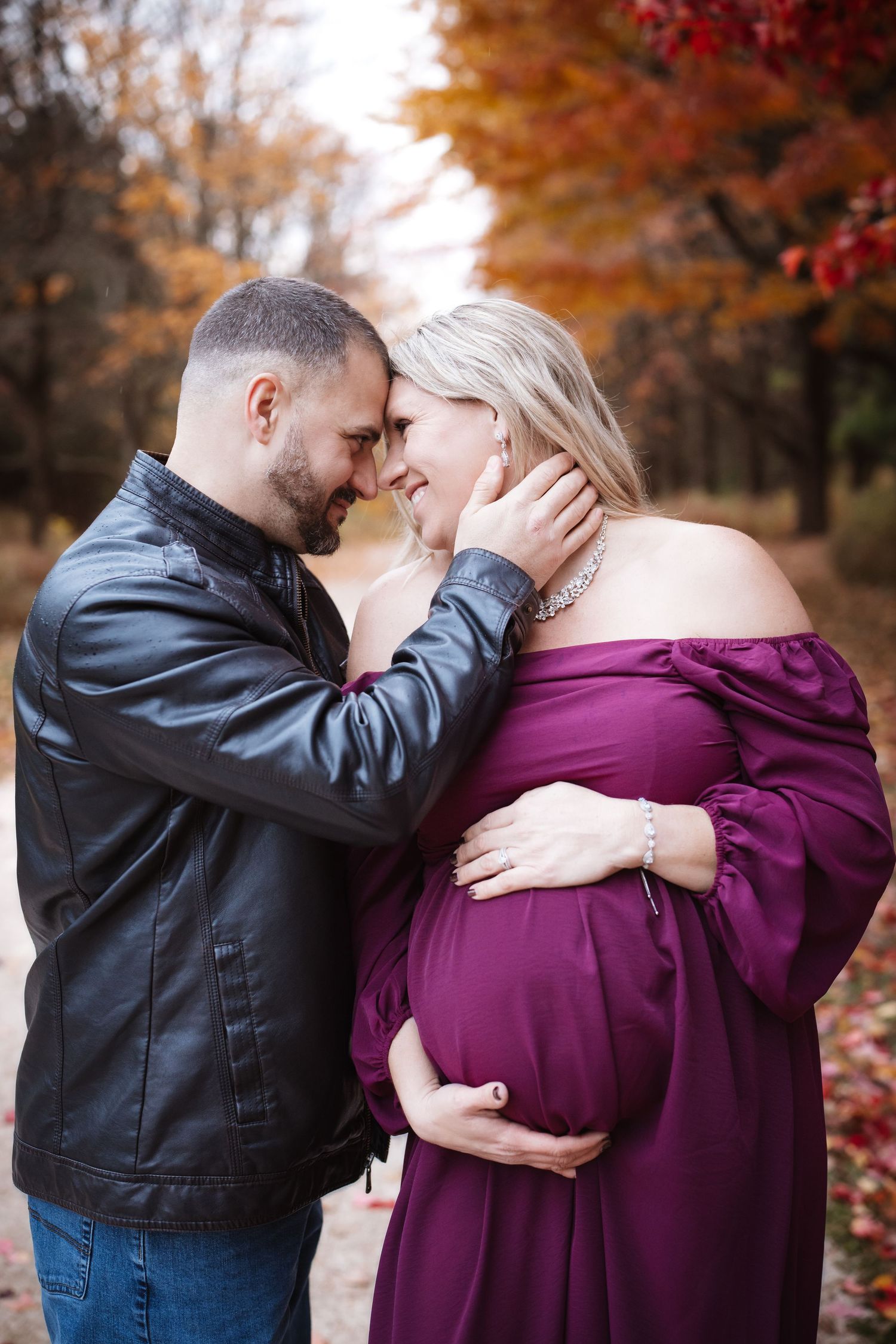 Expectant couple touching foreheads romantically outdoors, man cradles womans face as she holds baby bump in purple dress.
