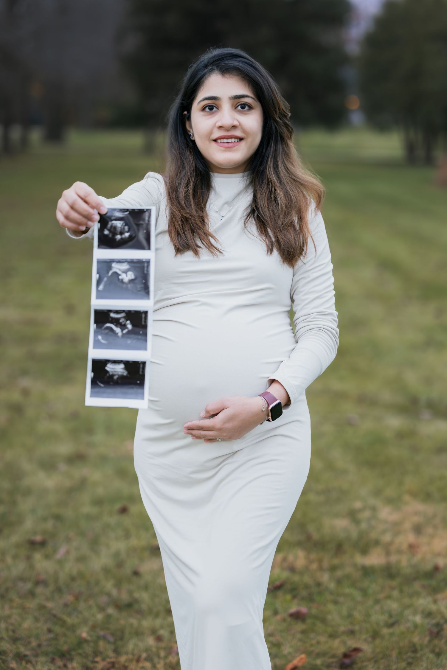 Pregnant woman in white dress smiling while holding ultrasound photos outdoors.