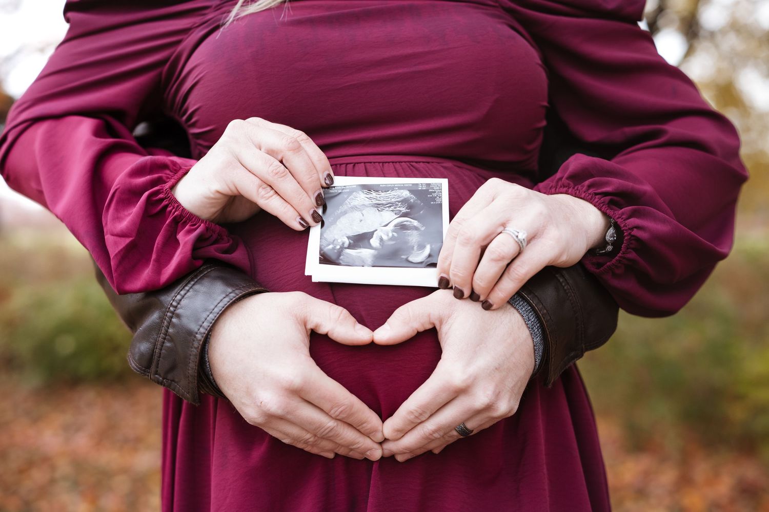 Couple holding ultrasound photo over pregnant belly making heart shape with hands outdoors.