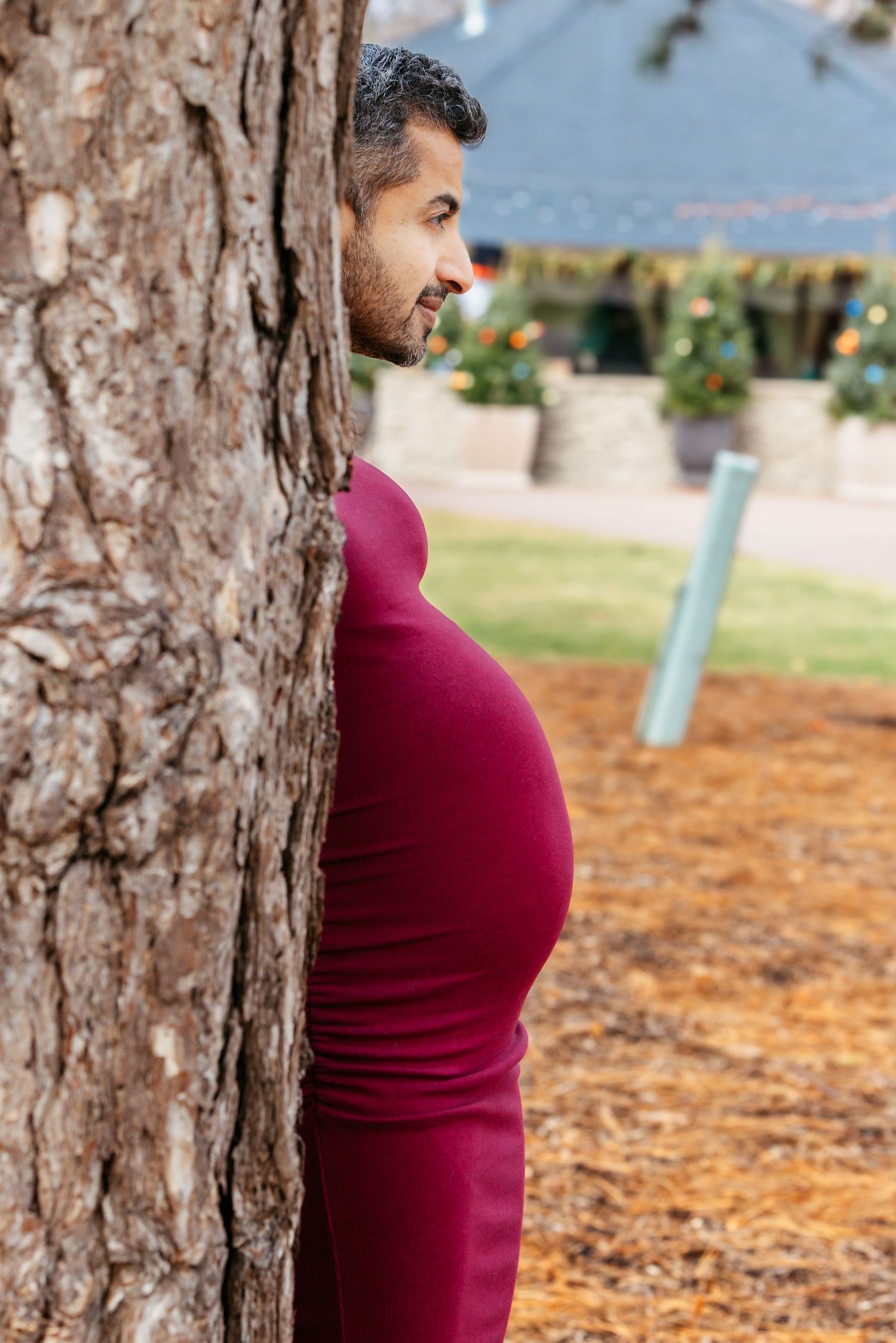 Pregnant woman in burgundy dress standing beside a large tree in a park.