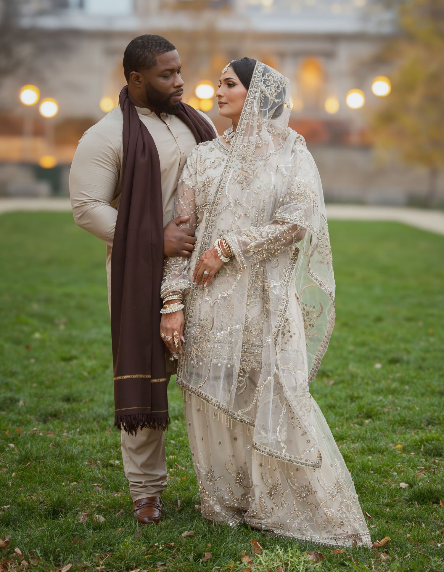 Couple in traditional South Asian wedding outfits share a tender moment on a green lawn at dusk.