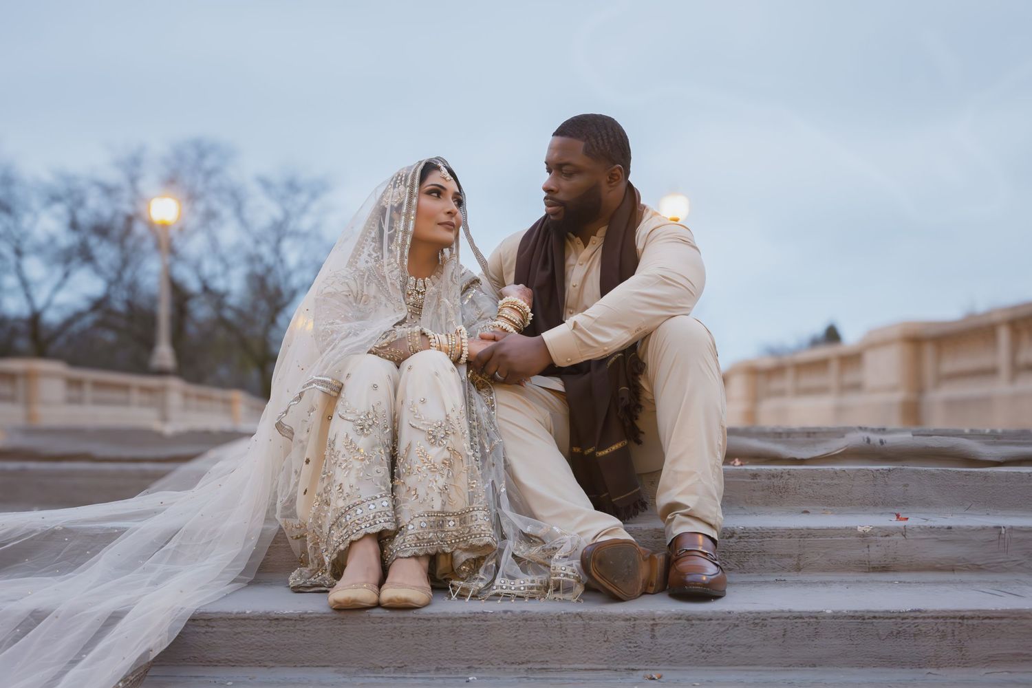 South Asian bride in gold embroidered lehenga and groom in cream sherwani sit on steps at dusk.