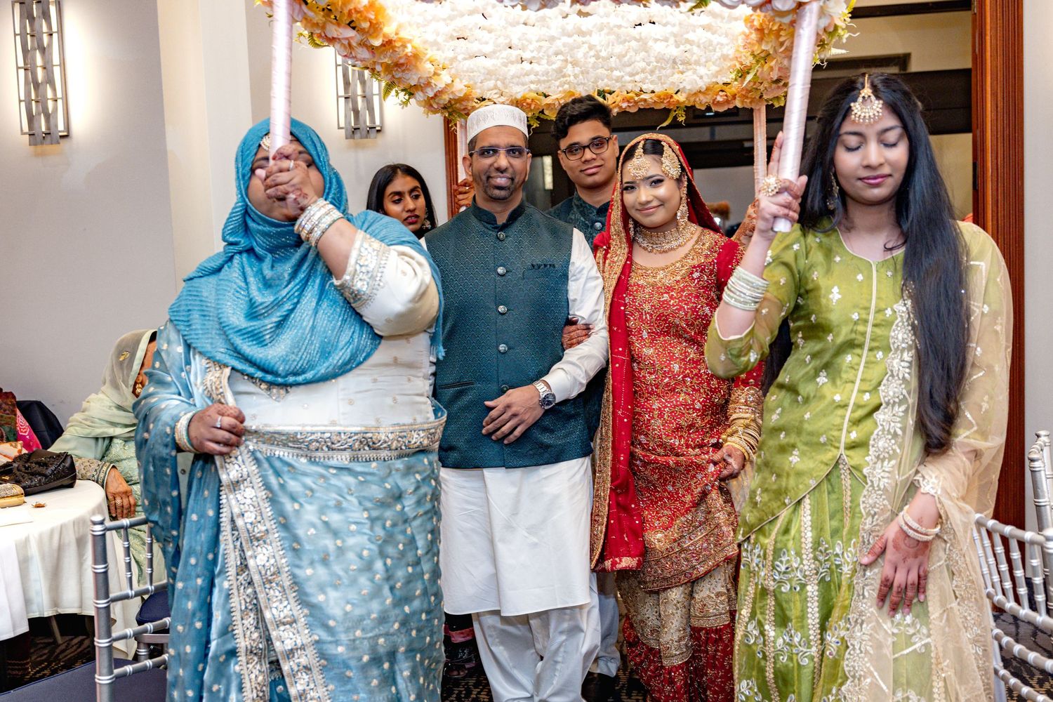 South Asian wedding party stands under floral canopy during traditional bride entry ceremony indoors.