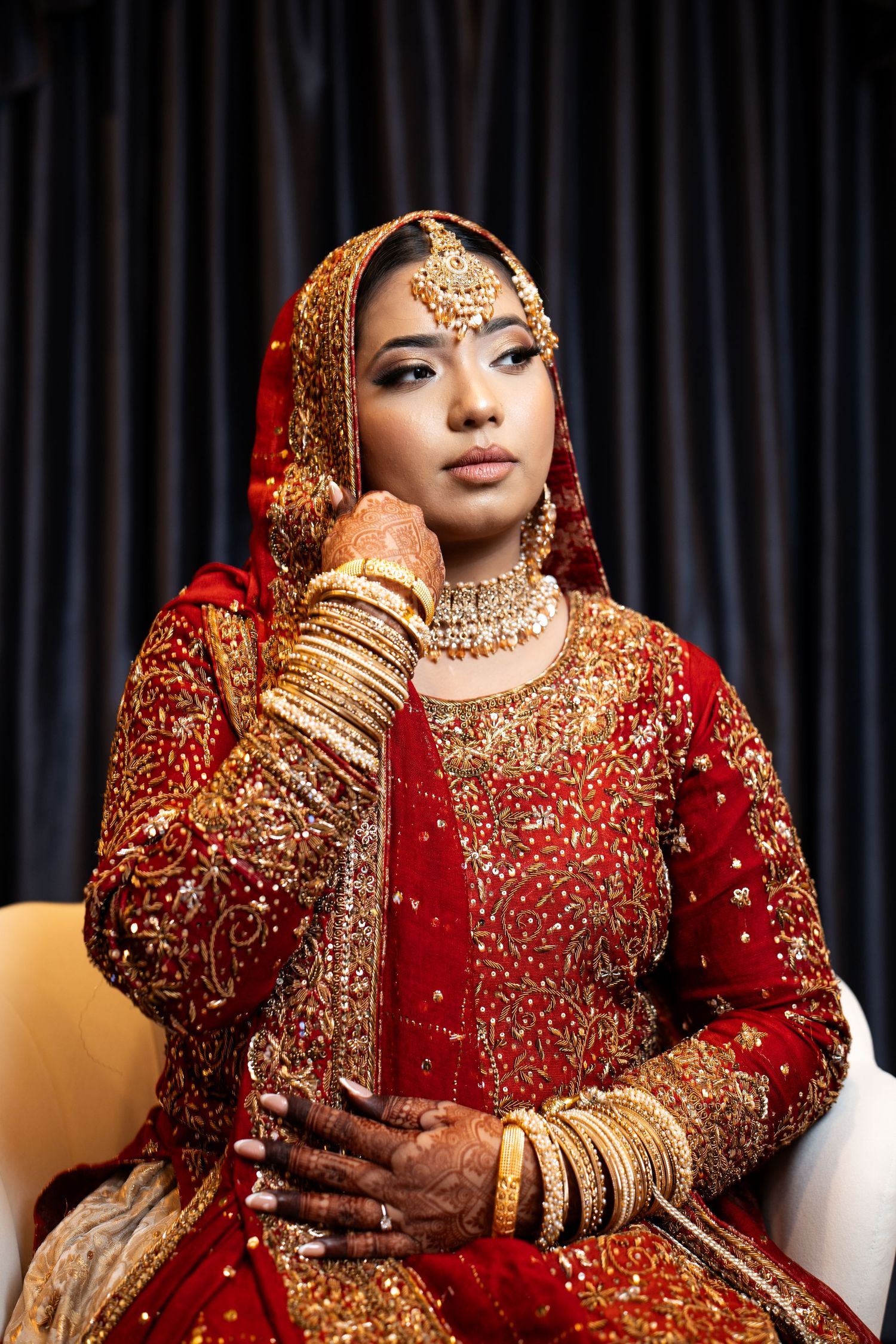 Bride in red and gold traditional South Asian bridal attire with ornate jewelry poses seated.