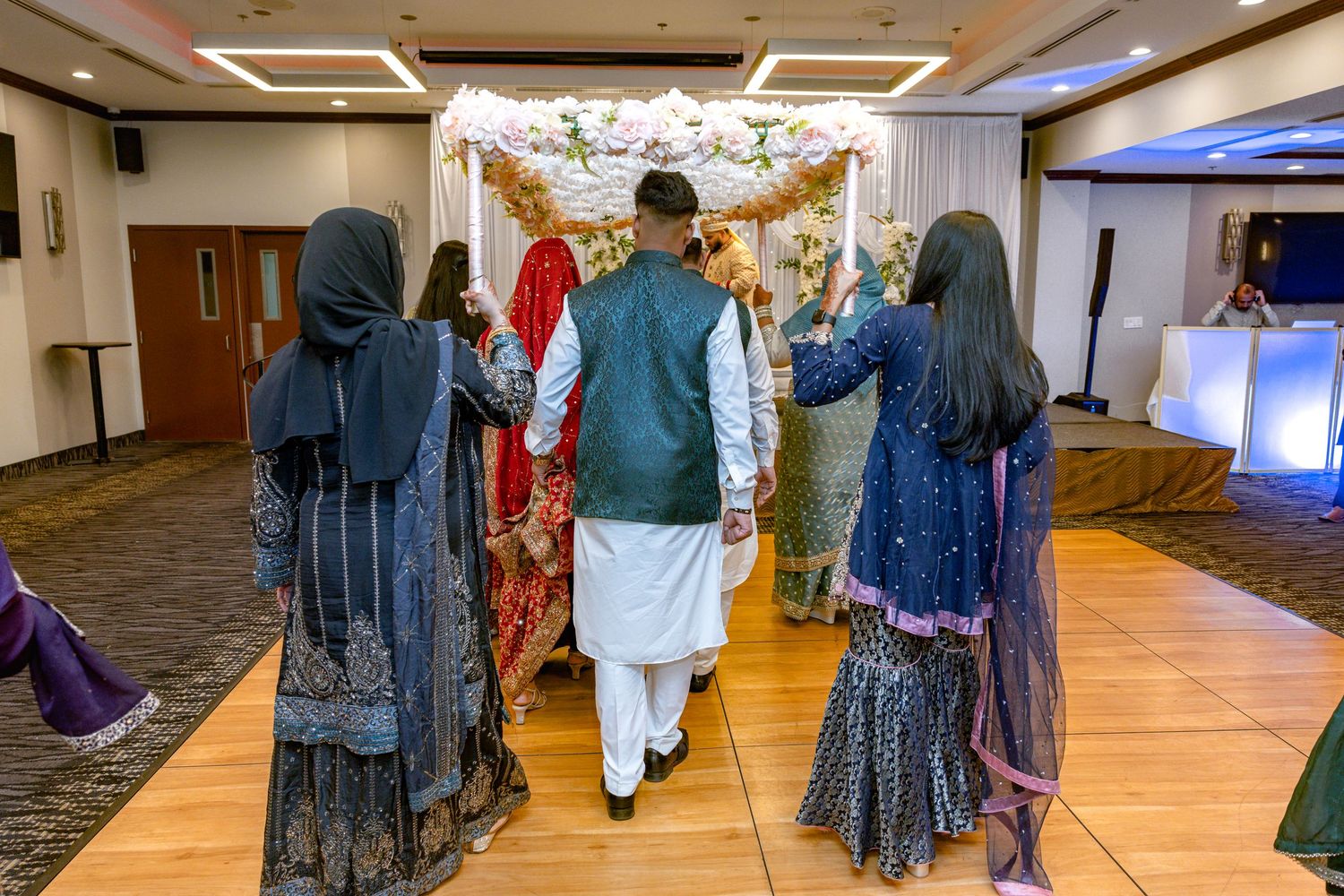 South Asian wedding procession with bride and groom walking under floral canopy held by family members.