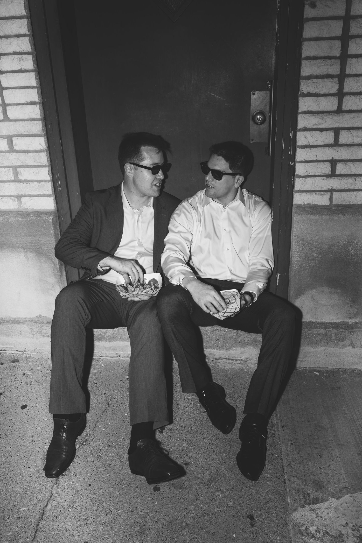 Black and white photo of two men in suits and sunglasses sitting on steps sharing a meal outdoors.