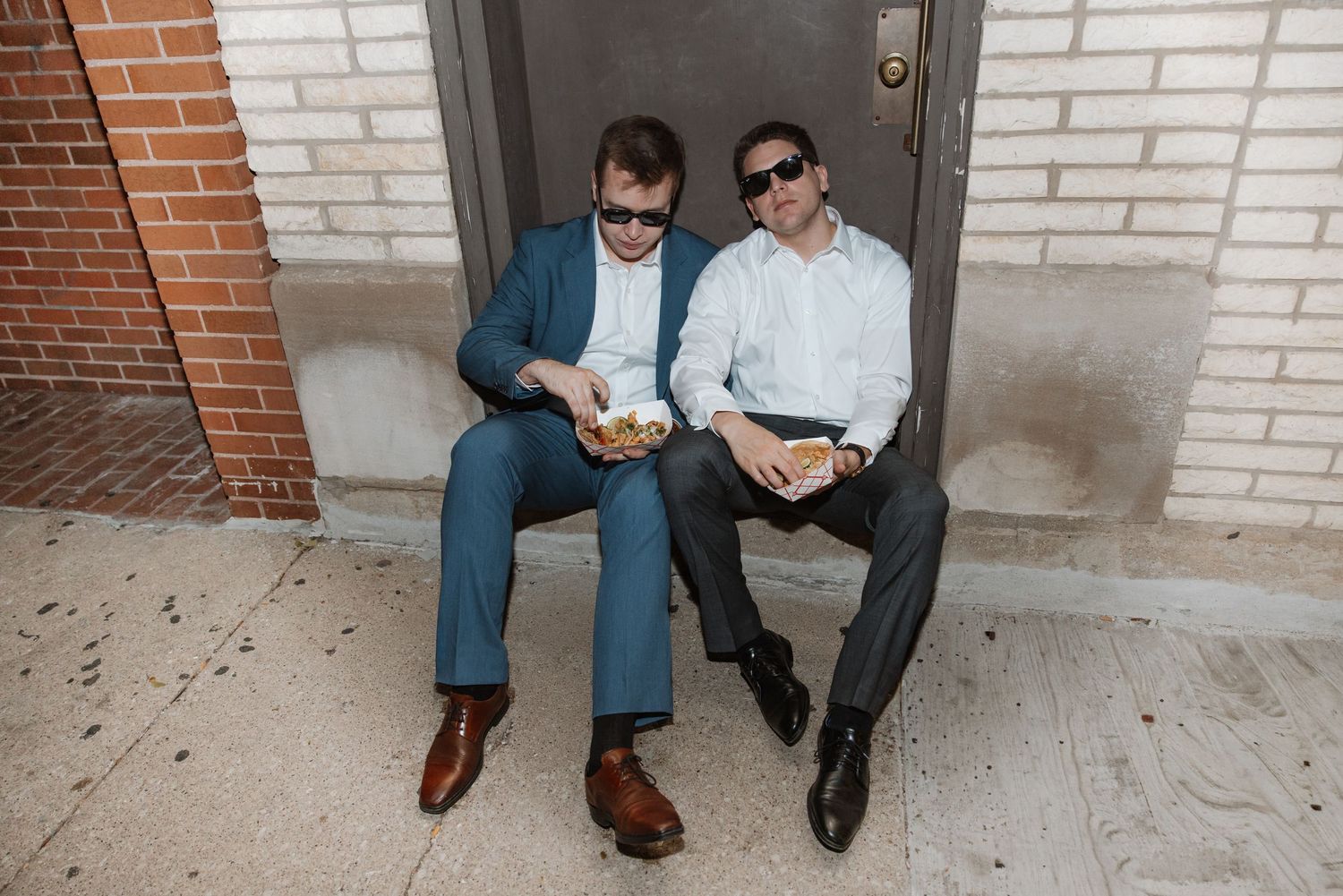 Two men in suits and sunglasses leaning together while eating food on outdoor steps.