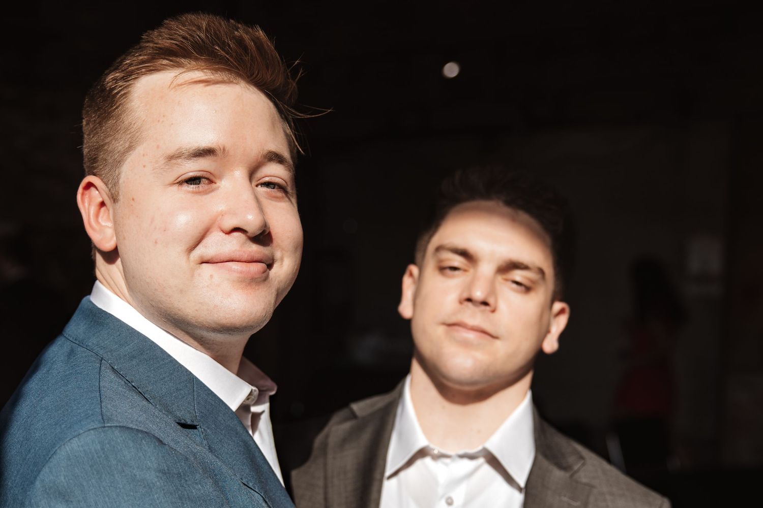 Close-up of two young men in suits smiling together against a dark background.