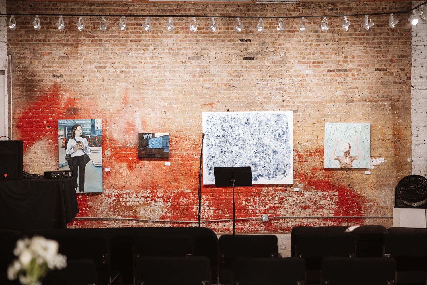 Exposed brick wall art gallery display with colorful paintings and a music stand in an industrial event space.