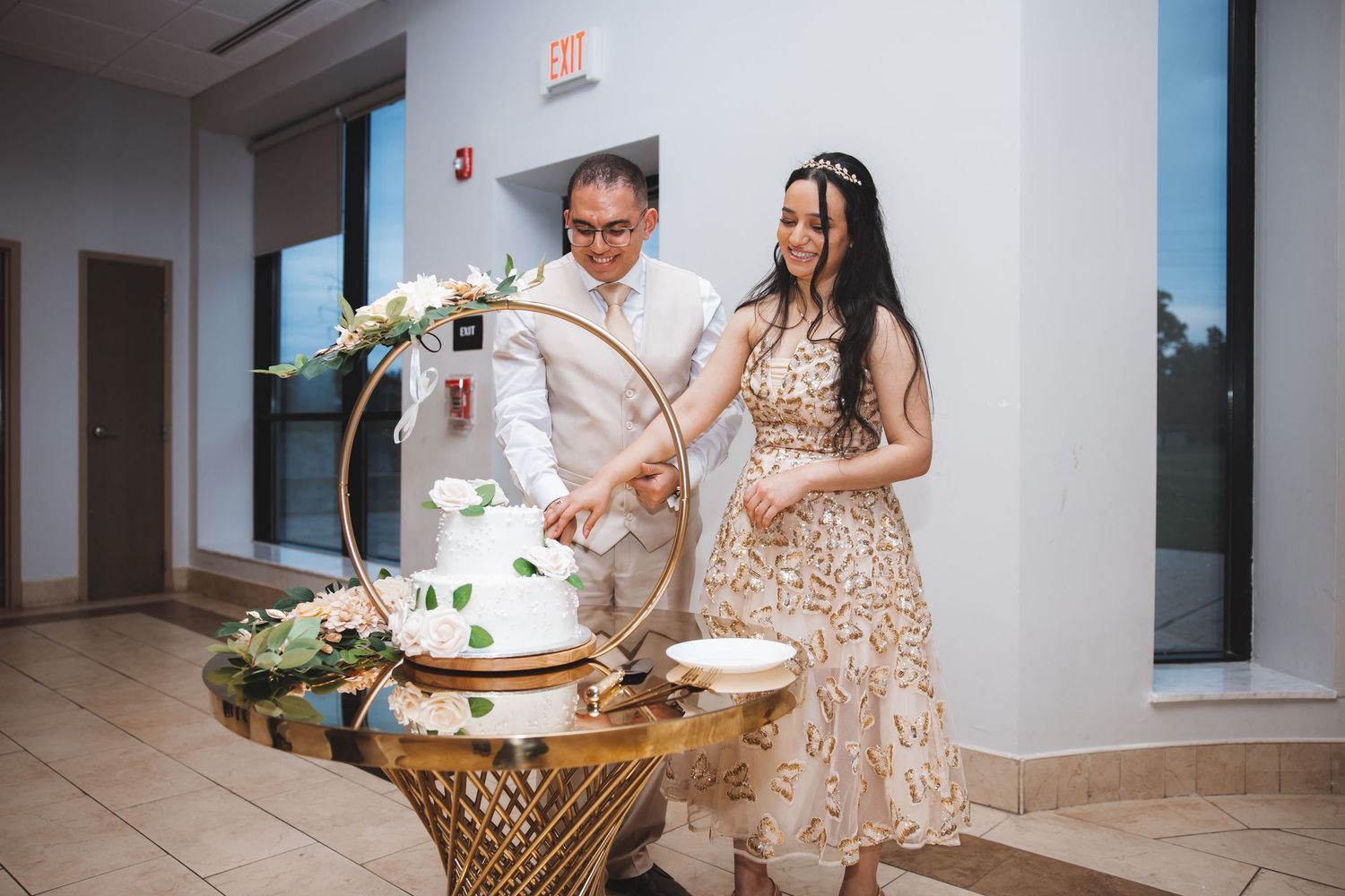 Newlyweds cutting white wedding cake on gold hoop stand at intimate reception.