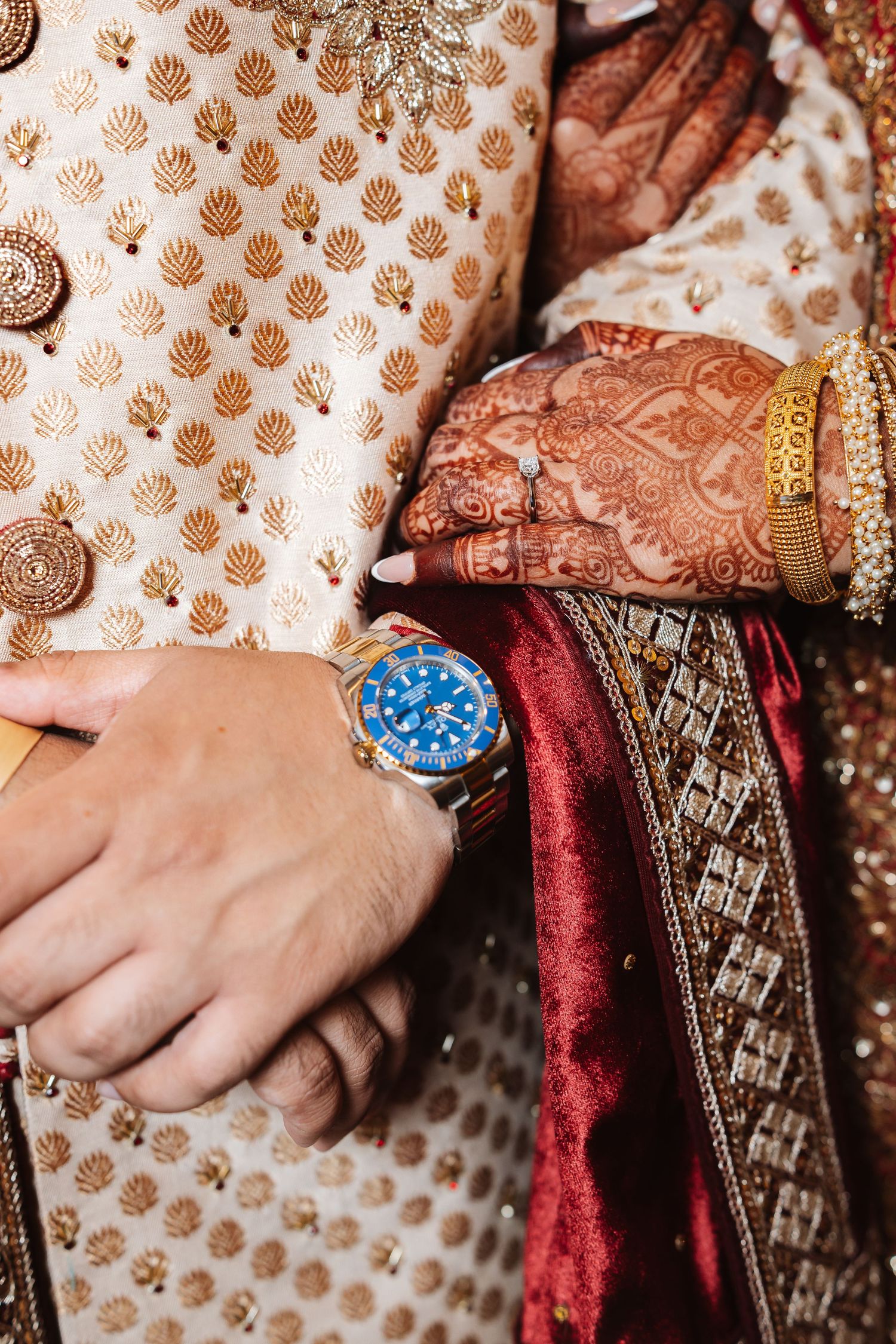 Close-up of Indian wedding couple, groom wearing blue Rolex watch, bride with mehndi and gold bangles.