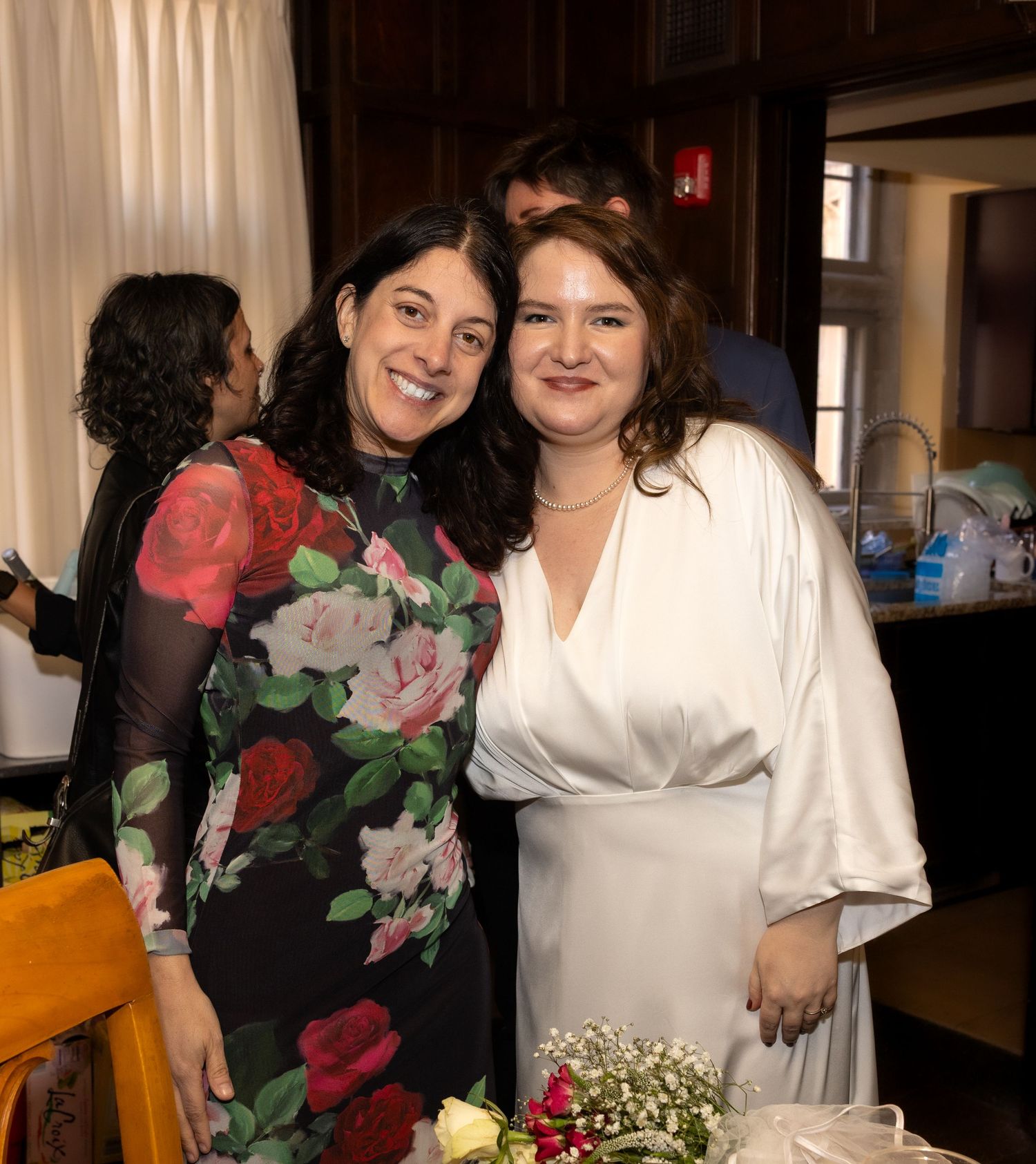 Two women smile together at a wedding reception, one wearing a white satin bridal dress.