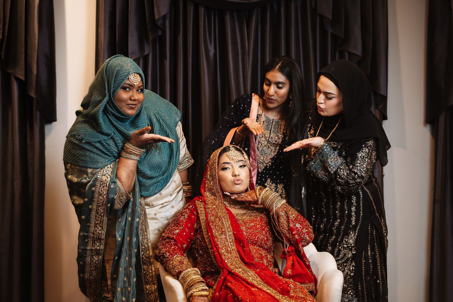 South Asian bride in red lehenga seated while three women in hijabs style her hair and makeup.