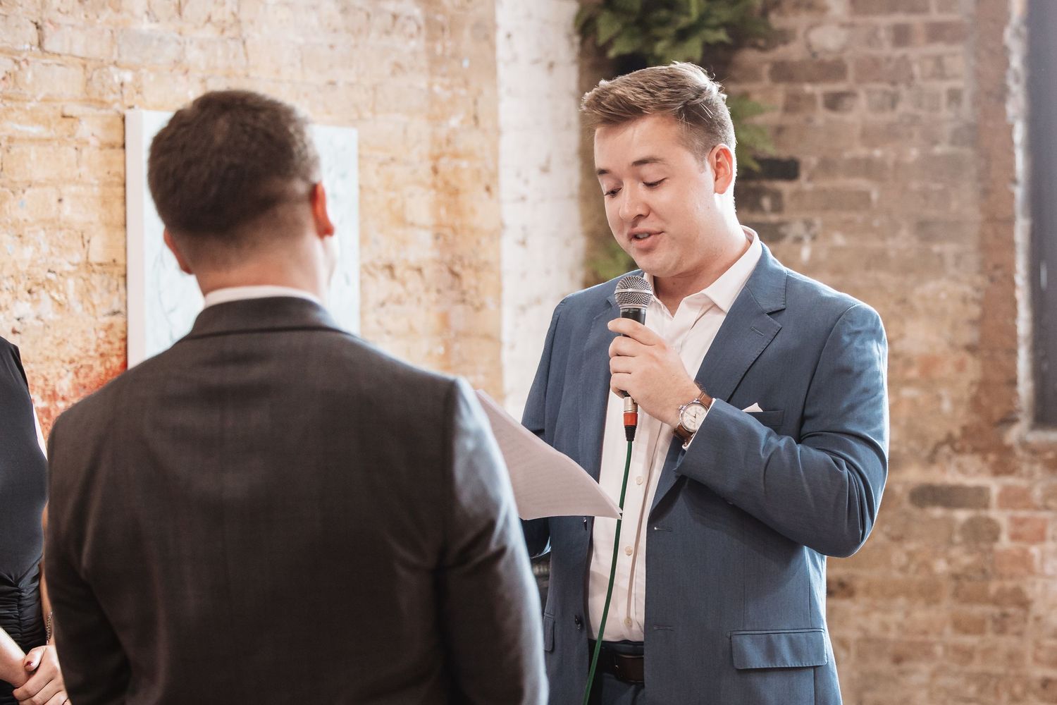 Young man in blue suit reading from paper while speaking into microphone at wedding ceremony.