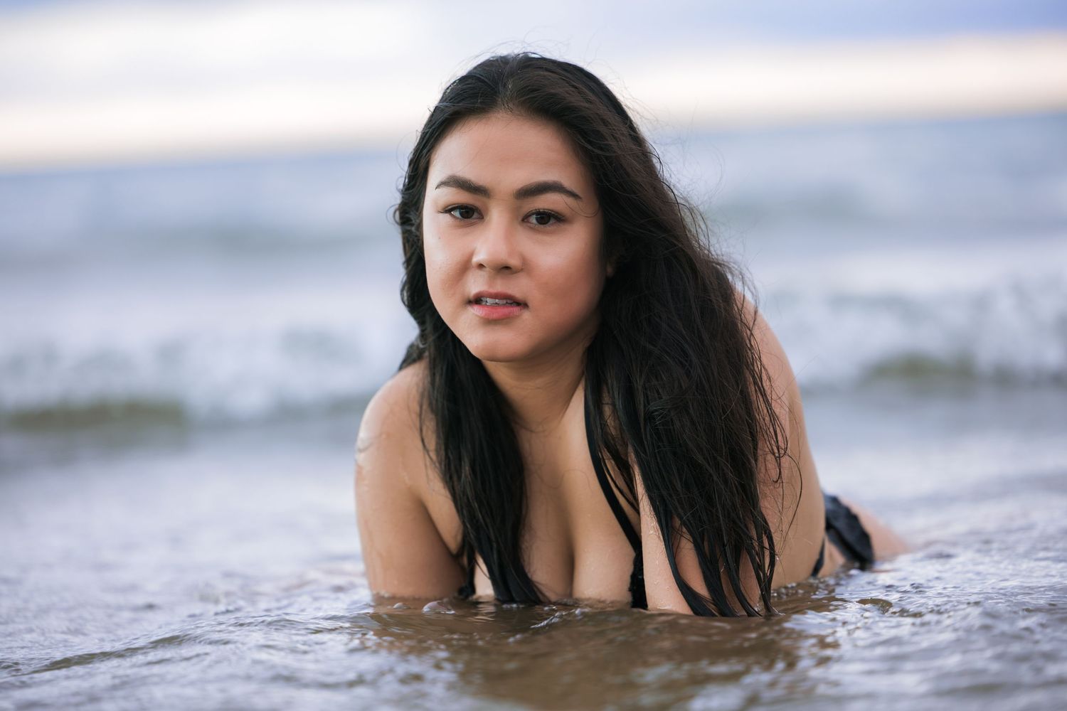 Young woman in black bikini lying in shallow ocean waves looking at camera on cloudy day.