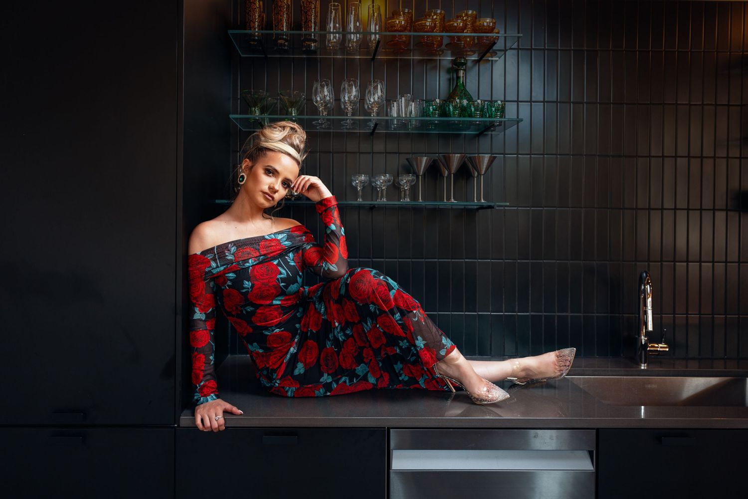 Woman in red floral off-shoulder dress sitting on dark bar counter barefoot.