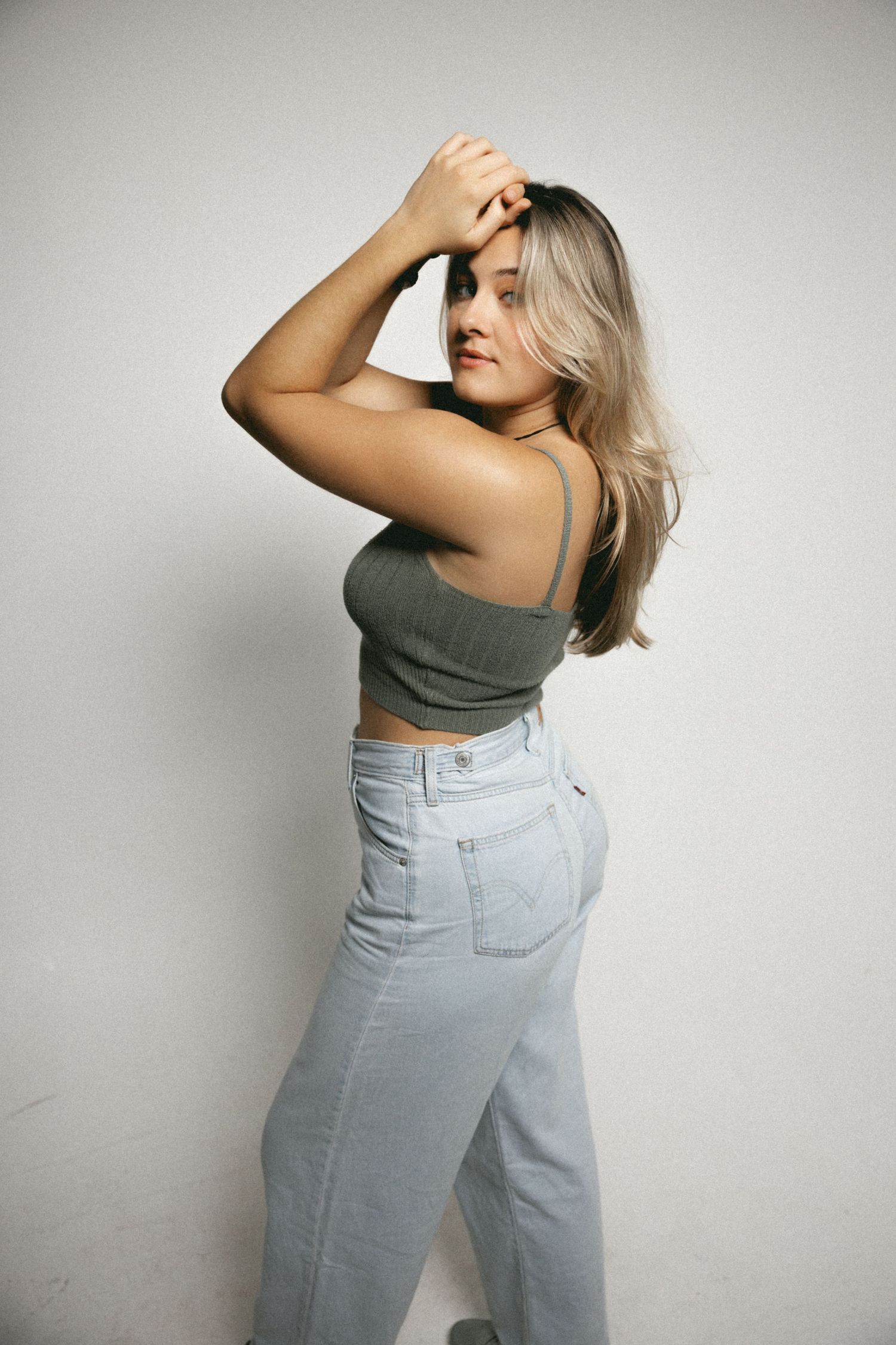 Blonde woman in olive crop top and high-waist light jeans poses sideways against white studio wall.