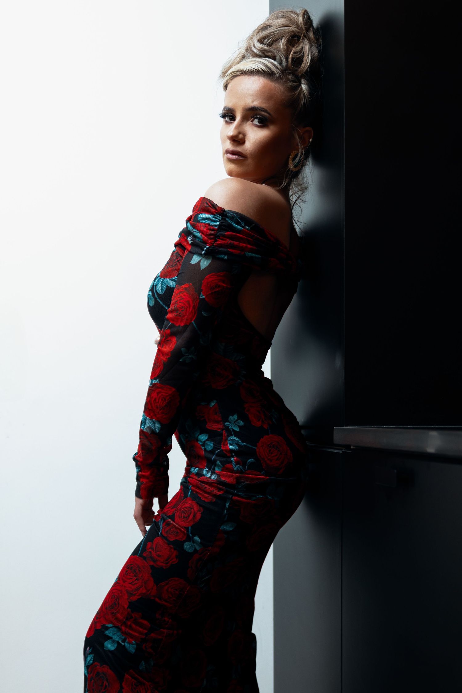 Woman in off-shoulder red rose print bodycon dress leaning elegantly against dark wall.