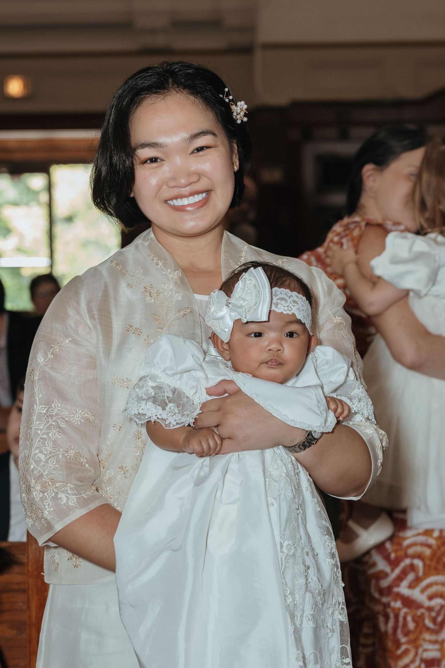 Smiling woman in white barong holds infant in christening gown at baptism.