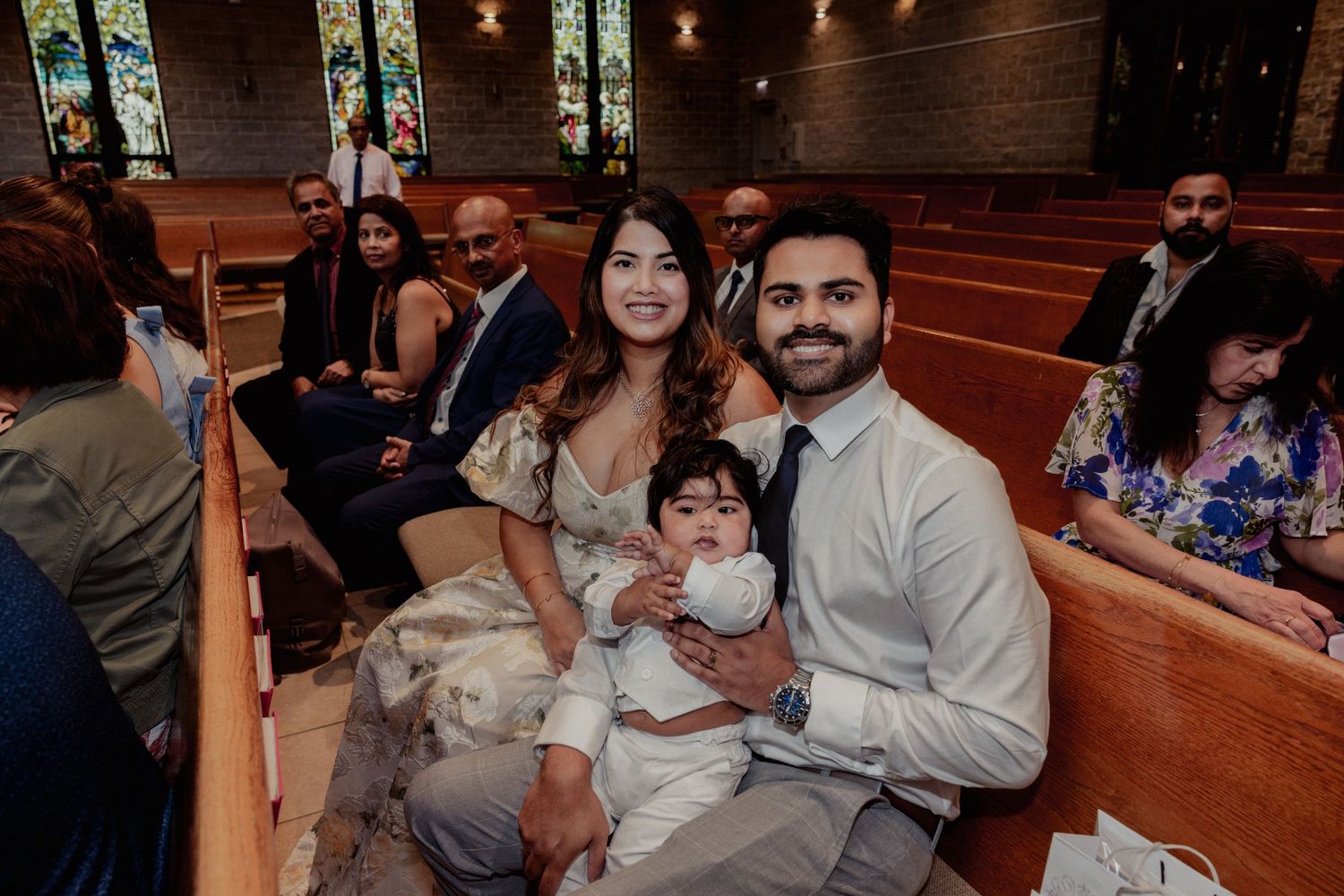 Family seated in church pew with baby in white christening outfit during baptism service.