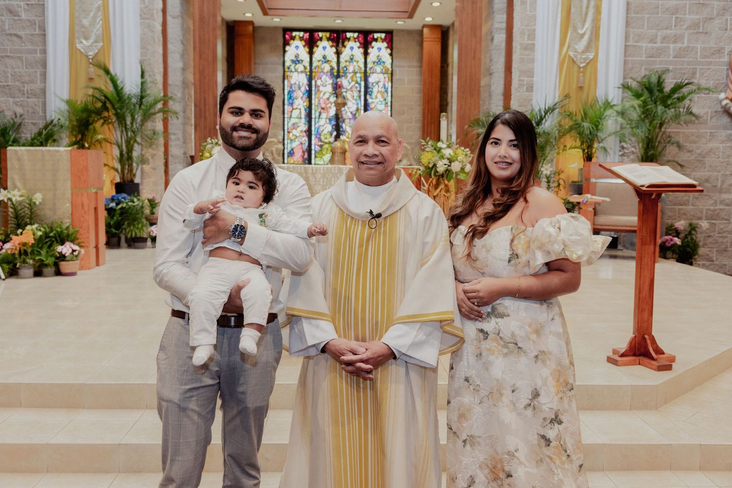 Family poses with Catholic priest after baptism ceremony in decorated church.