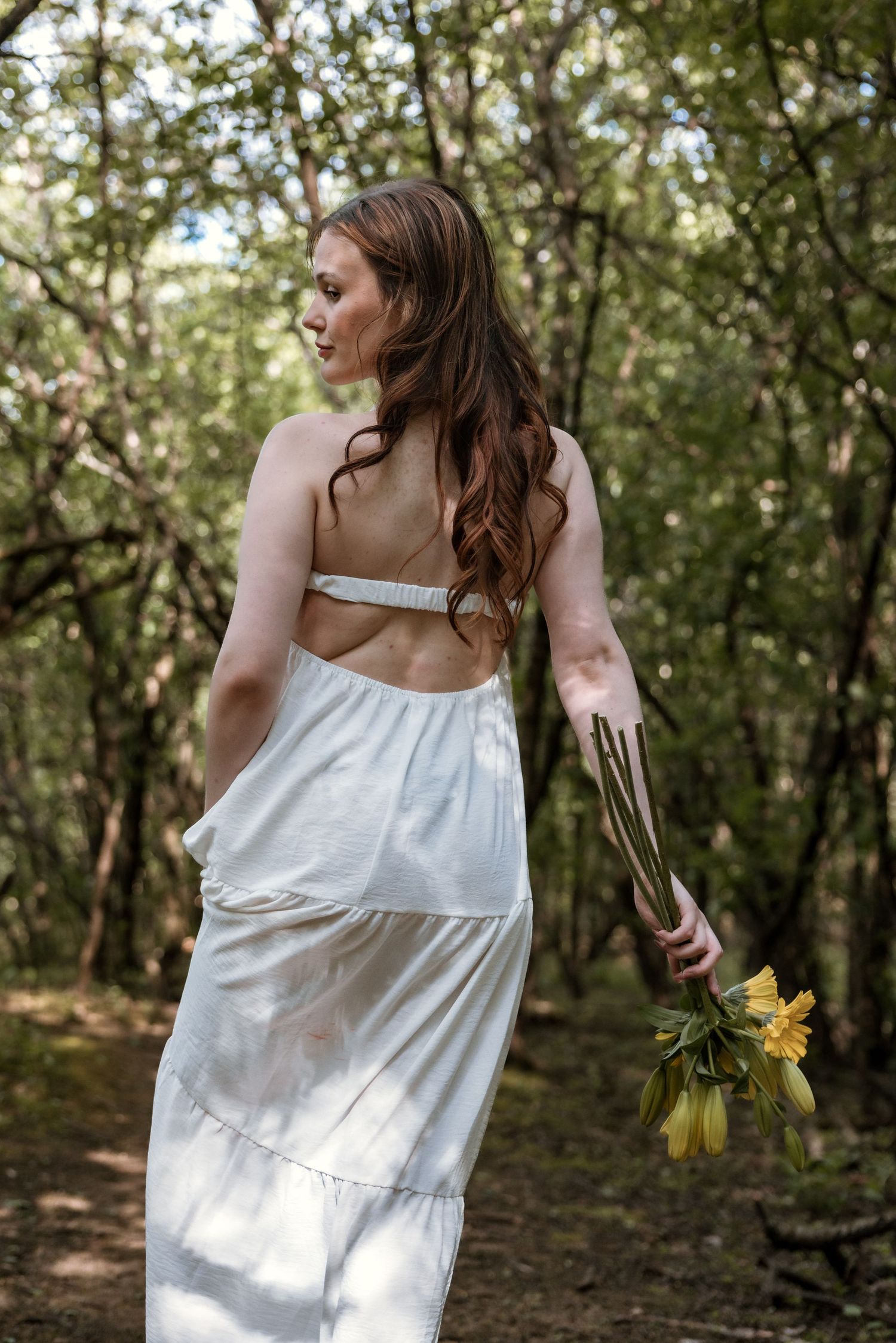 Young woman in open-back white dress holds yellow flowers while walking through woods.